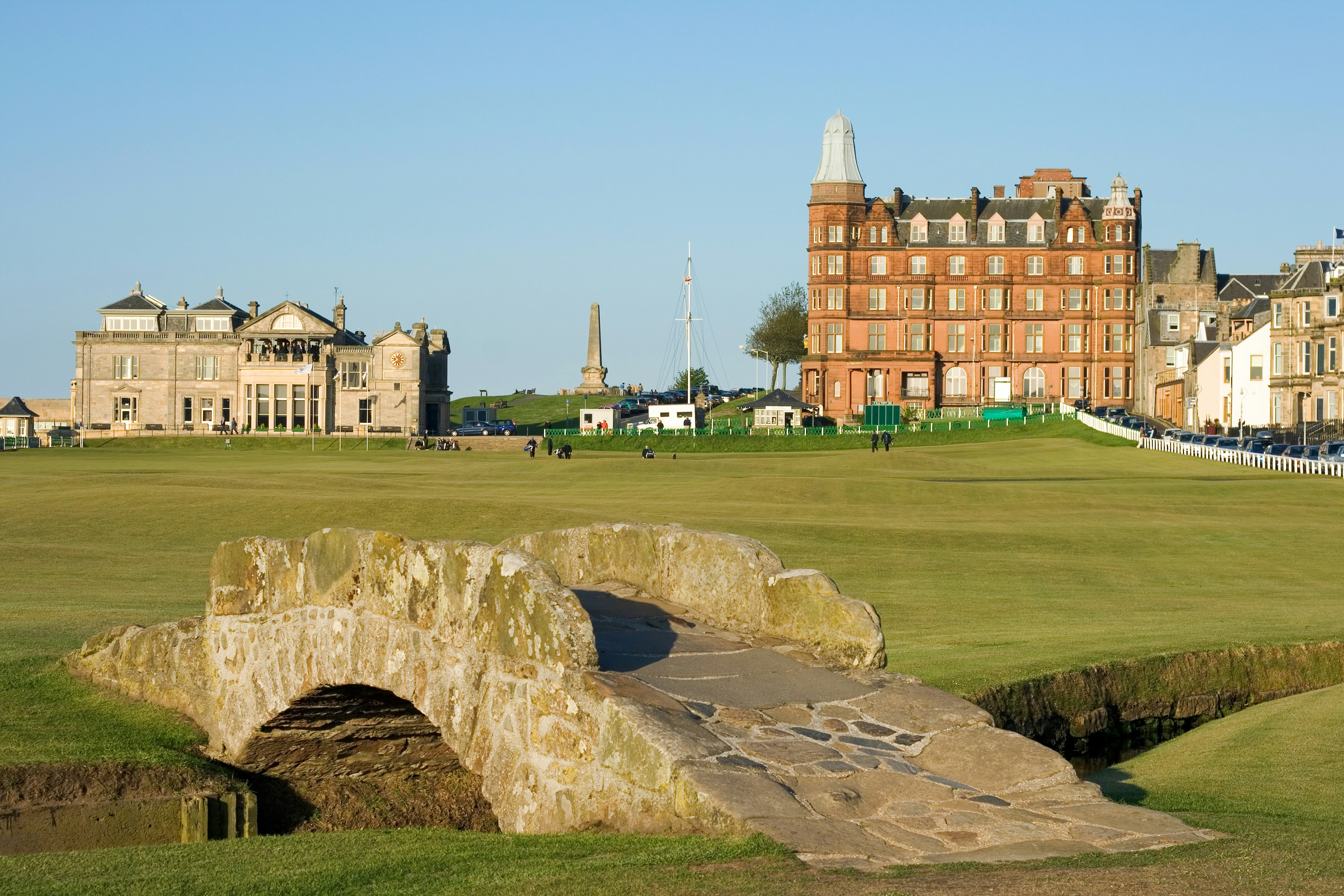 A stone bridge leads to the expansive greens of the St. Andrews Golf Course. In the background, there are a pair of old stone buildings. 