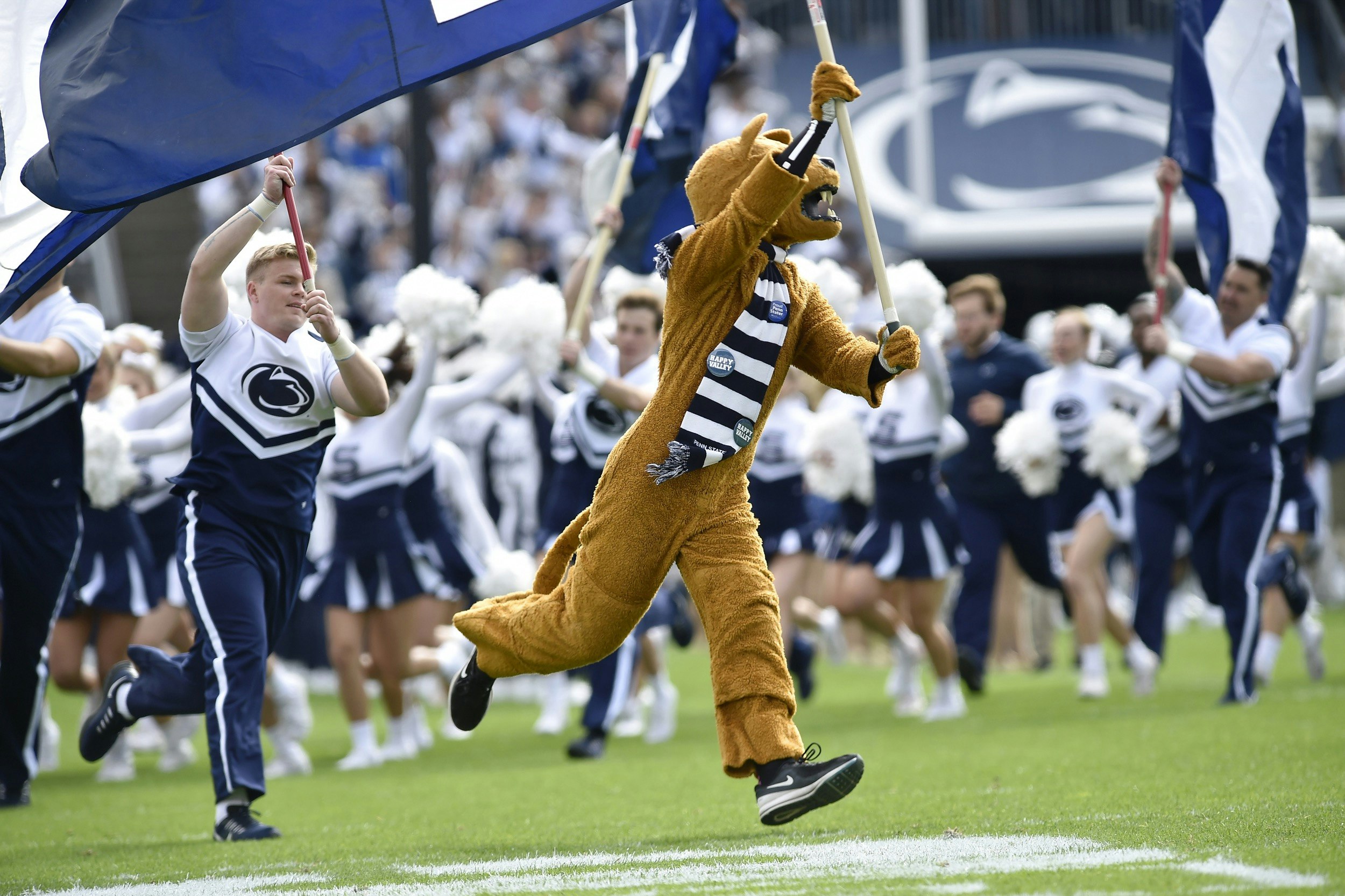The Nittany Lion Mascot, wearing a blue and white striped scarf, leads the Penn State Cheerleaders onto the field as he or she waves a flag with the school's football logo; Fall in State College Pennsylvania