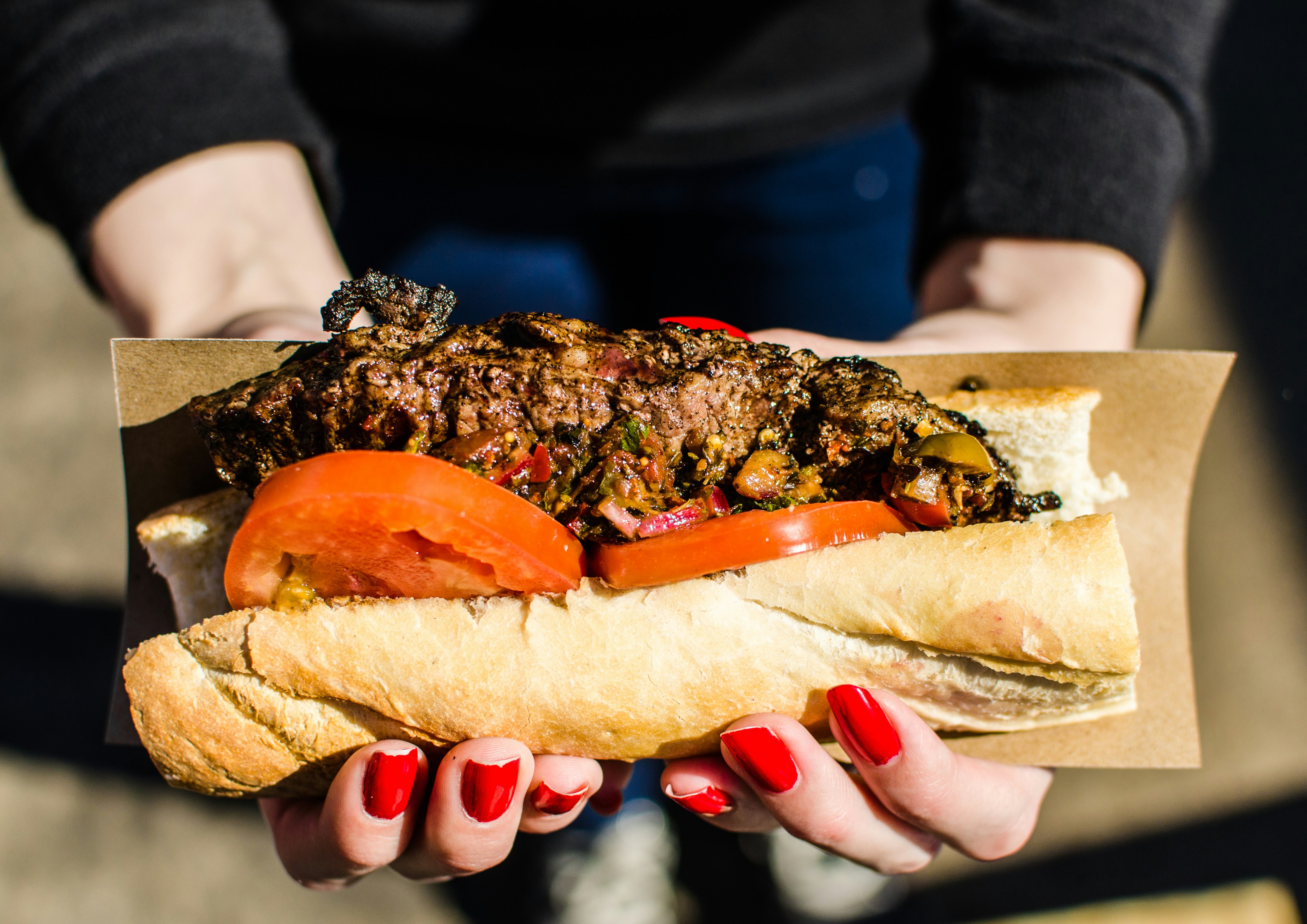 A woman with bright red nails holds a large steak sandwich. Two tomatoes are wedged on the side of the sandwich.