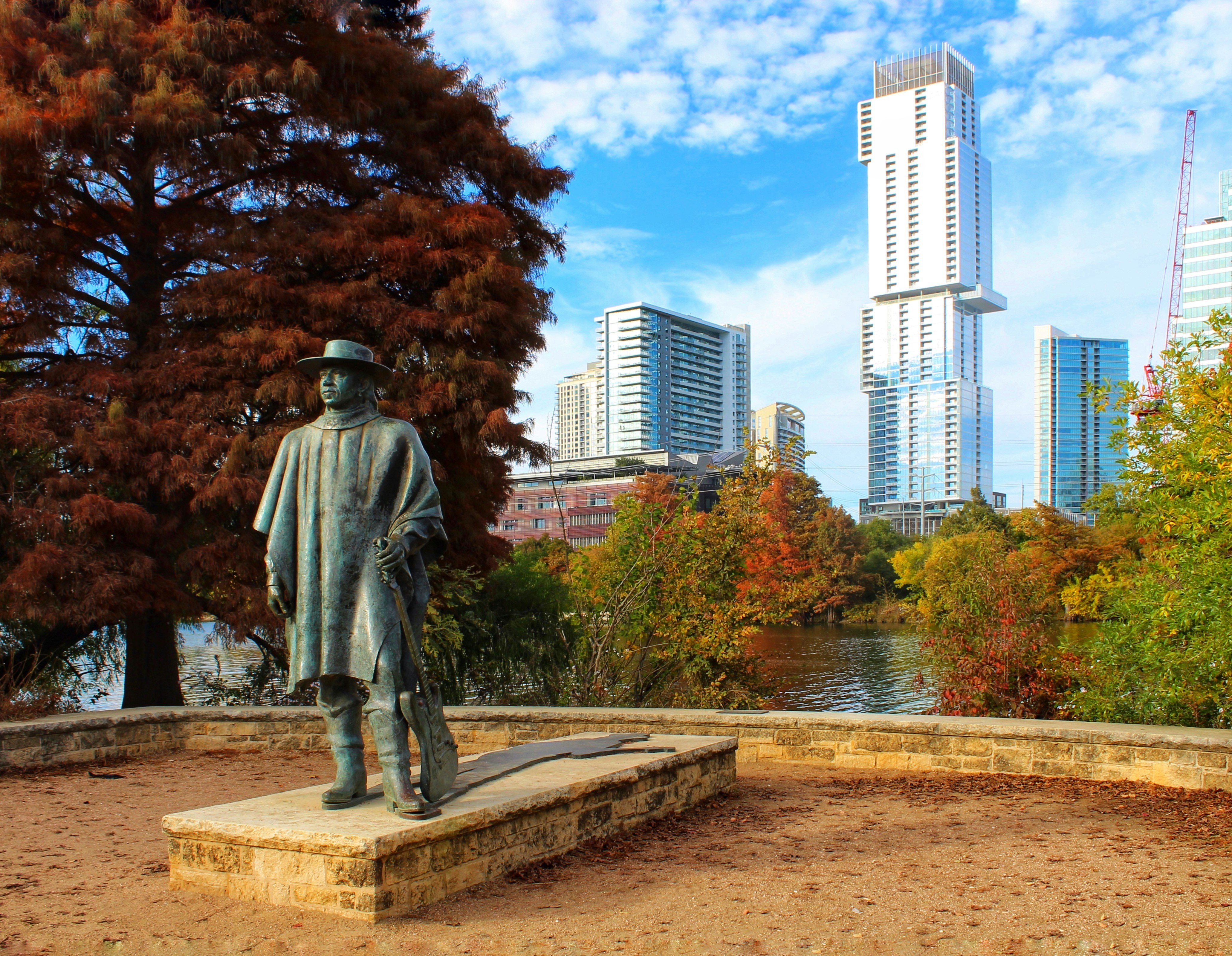 A statue of musician Stevie Ray Vaughan stands in Austin, Texas. The sun is shining and tall buildings are visible in the background. 