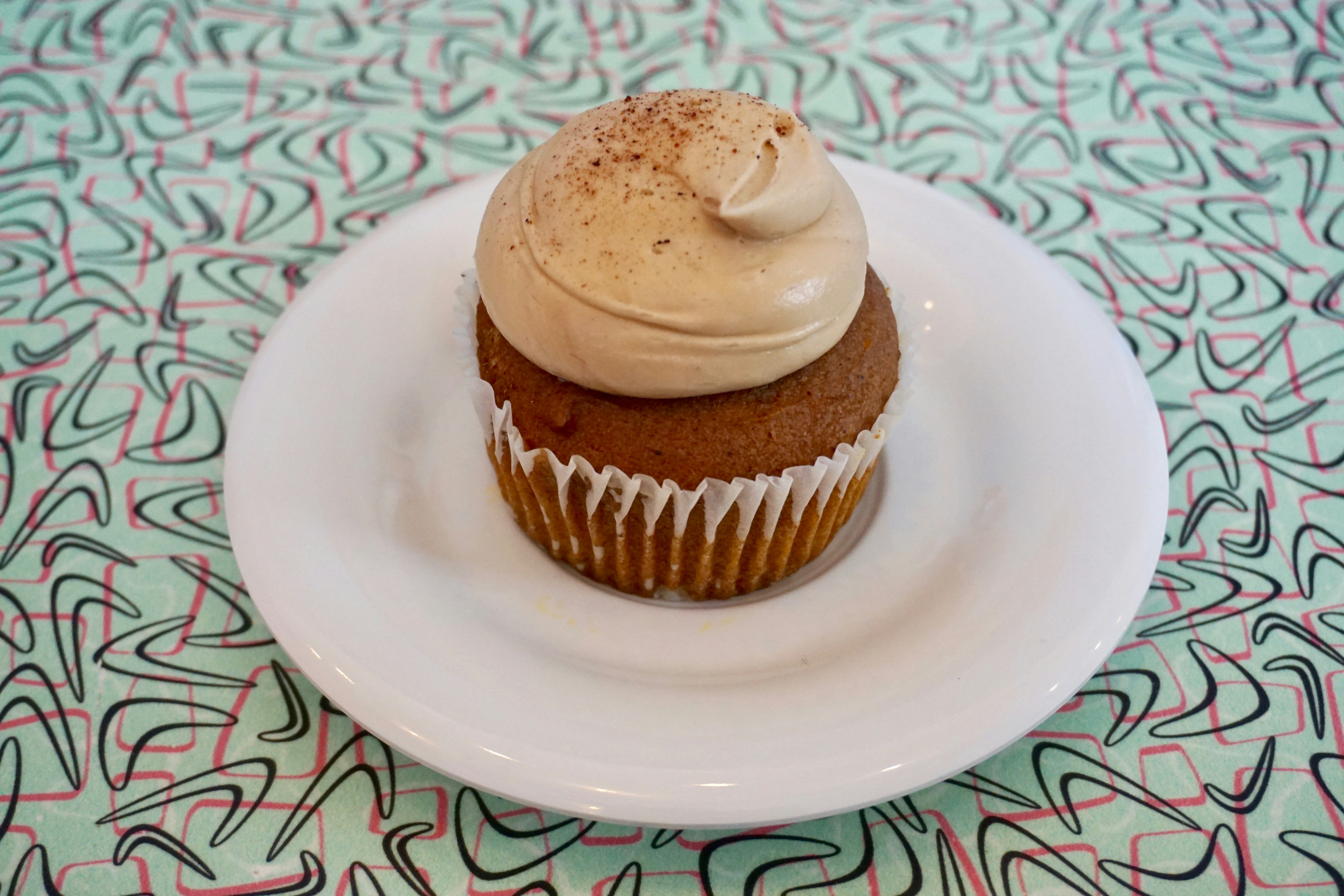 A cupcake with a generous swirl of icing sits on a white plate; underneath is a light blue tablecloth with a pink and black abstract pattern.