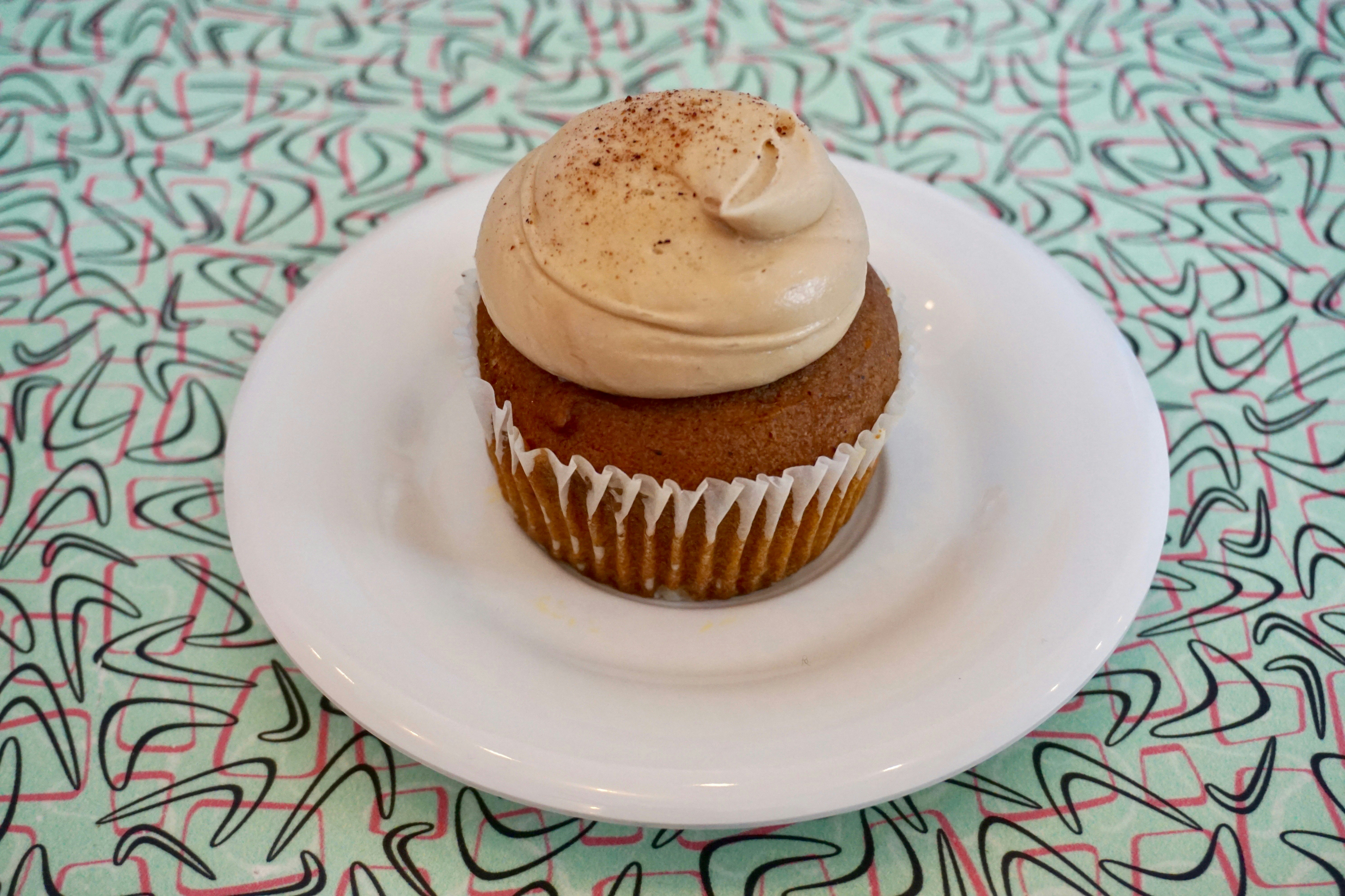 En cupcake med en generös virvel av glasyr sitter på en vit tallrik;  under är en ljusblå duk med rosa och svart abstrakt mönster.