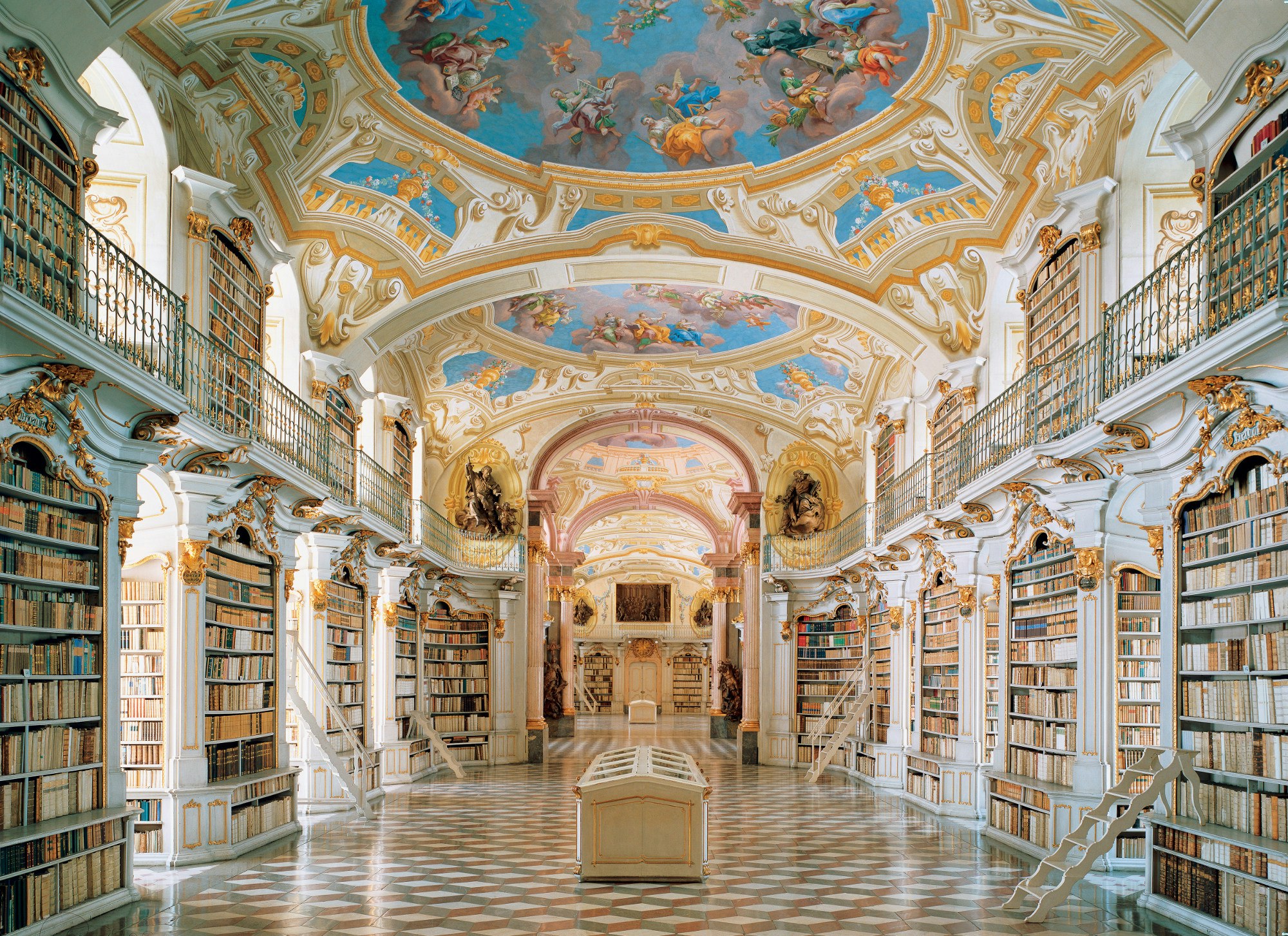 A long hall is supported by rectangular pillars that double as bookcases, each edged with white and gilt baroque columns. A balcony rings the upper level of the columns, and overhead a white, blue and gilt ceiling is made up of vaults and architectural flare.