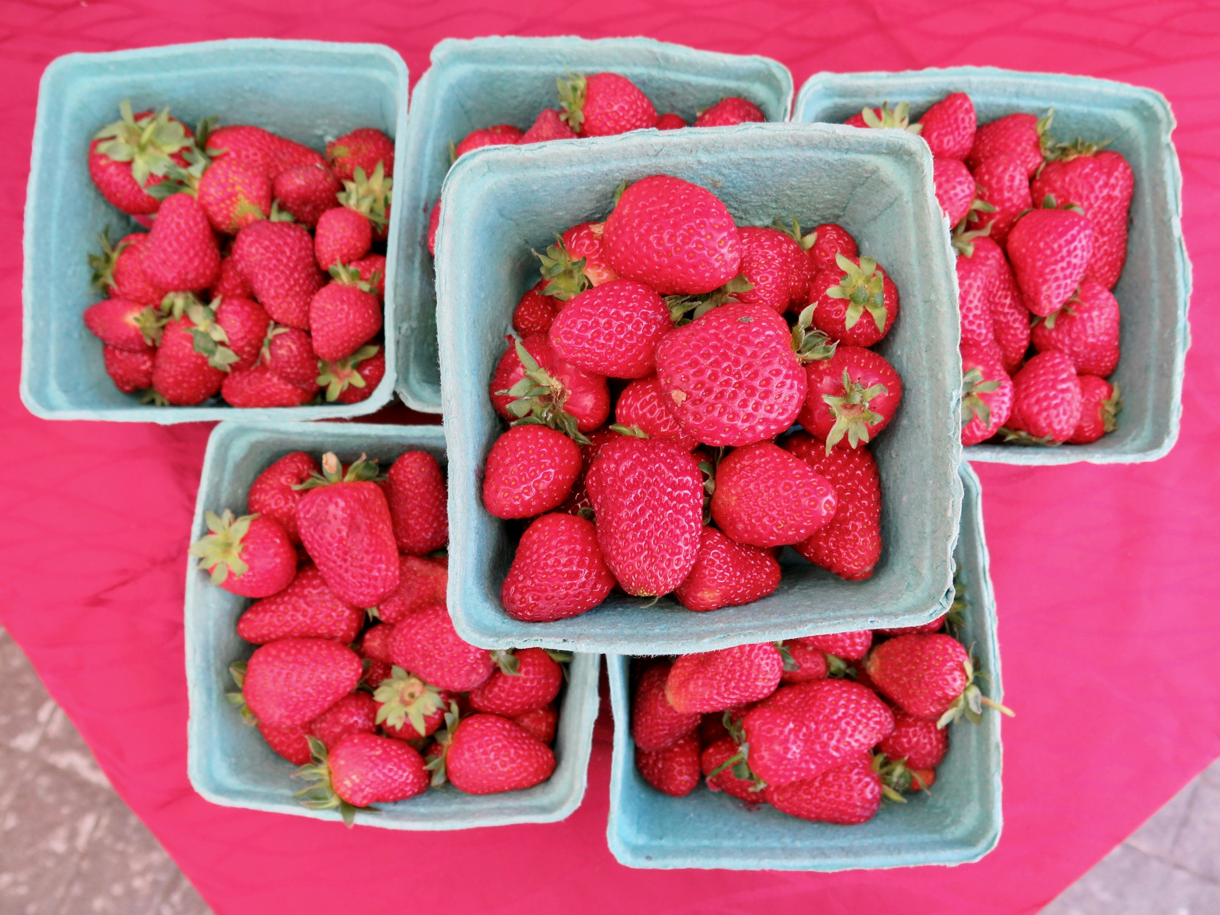 A top-down look at red strawberries sitting  in a small basket on a table with a red tablecloth.