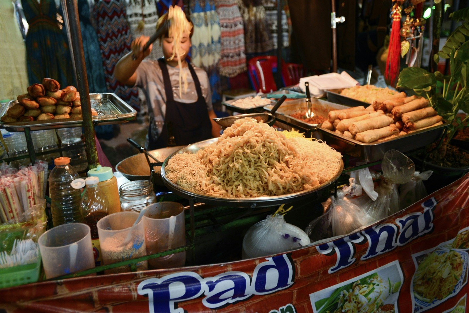 Woman stands behind a street food stand with a large wok filled with Pad Thai