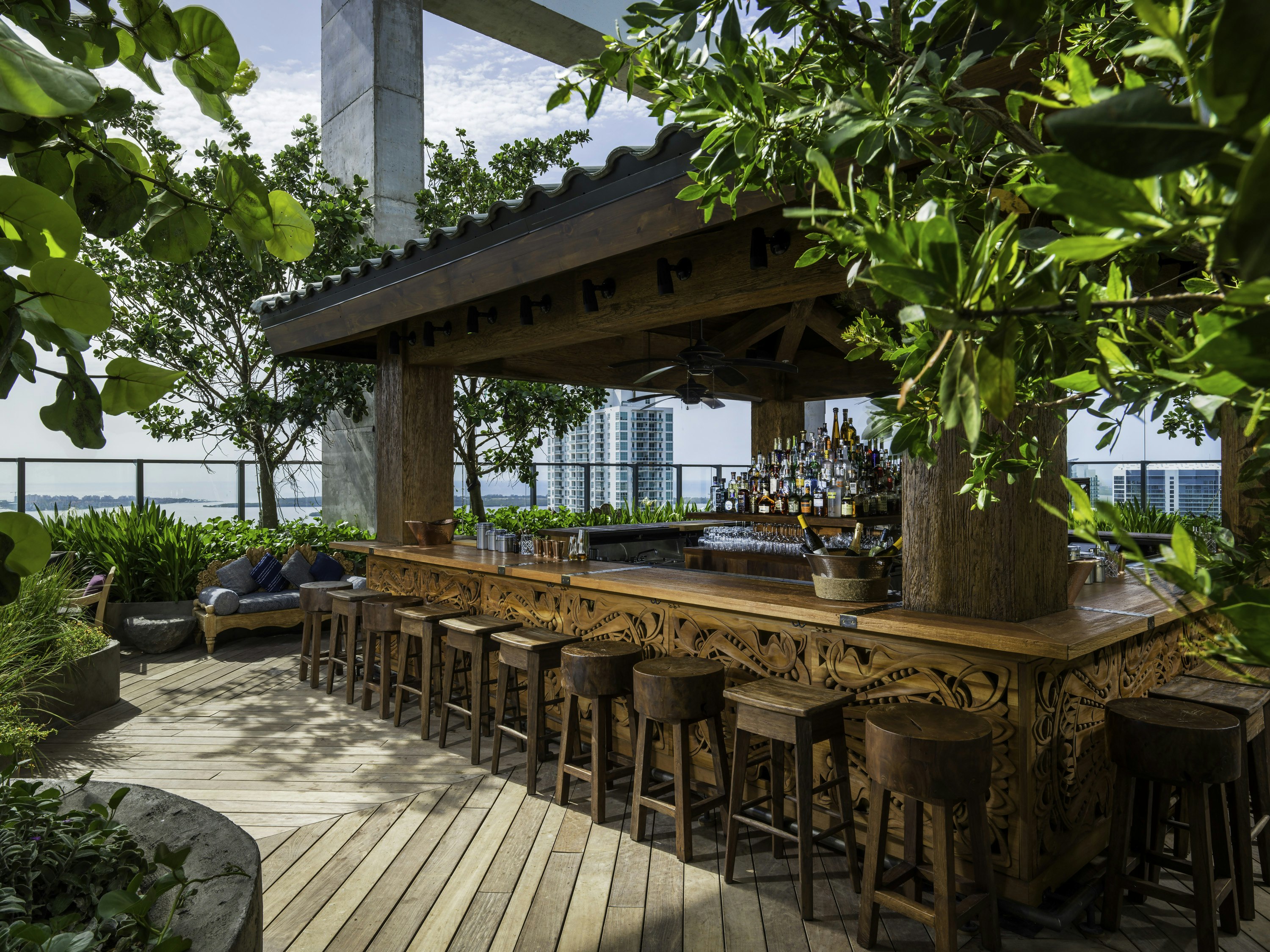A rooftop bar scene; in the centre there's a huge covered bar, with intricate wood-carvings and wooden bar stools lining it, and the area is decorated with lush greenery. Part of the Miami skyline is visible beyond glass railings.
