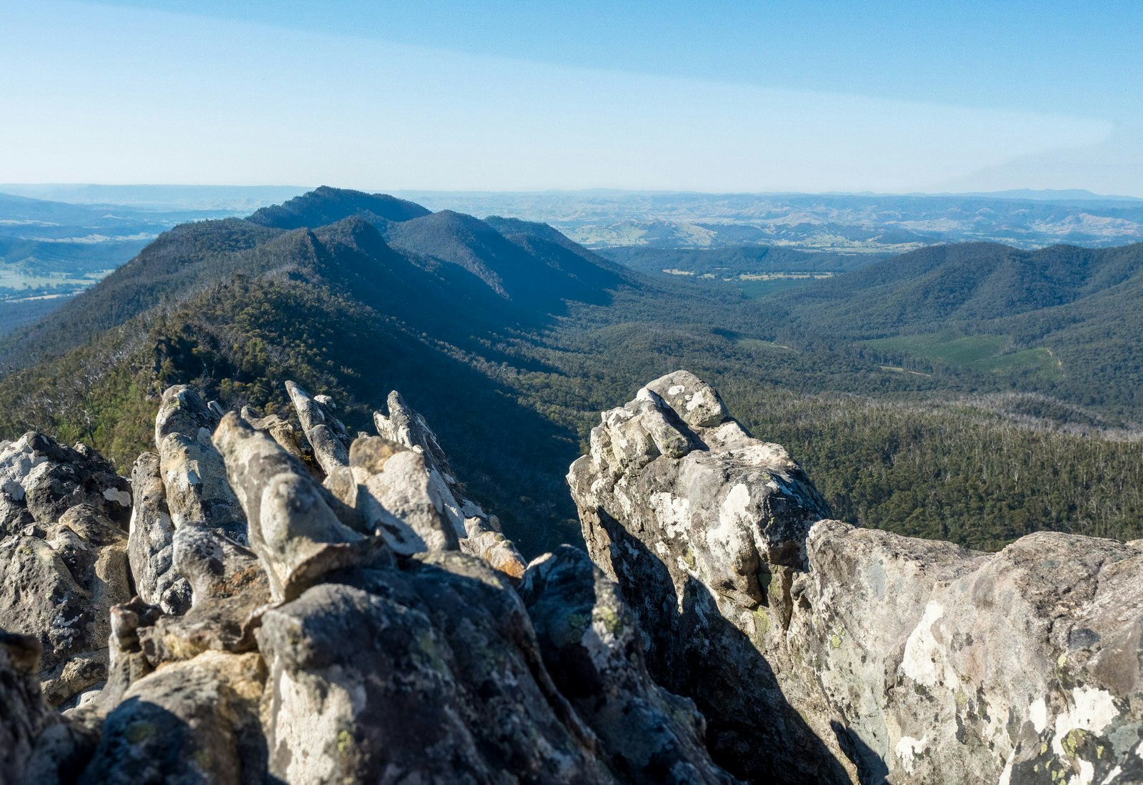 A panoramic shot of Cathedral Ranges State Park in Victoria. The sky is clear and blue and there are tree-covered hills all the way to the horizon. In the immediate foreground there is a worn formation of grey rock.