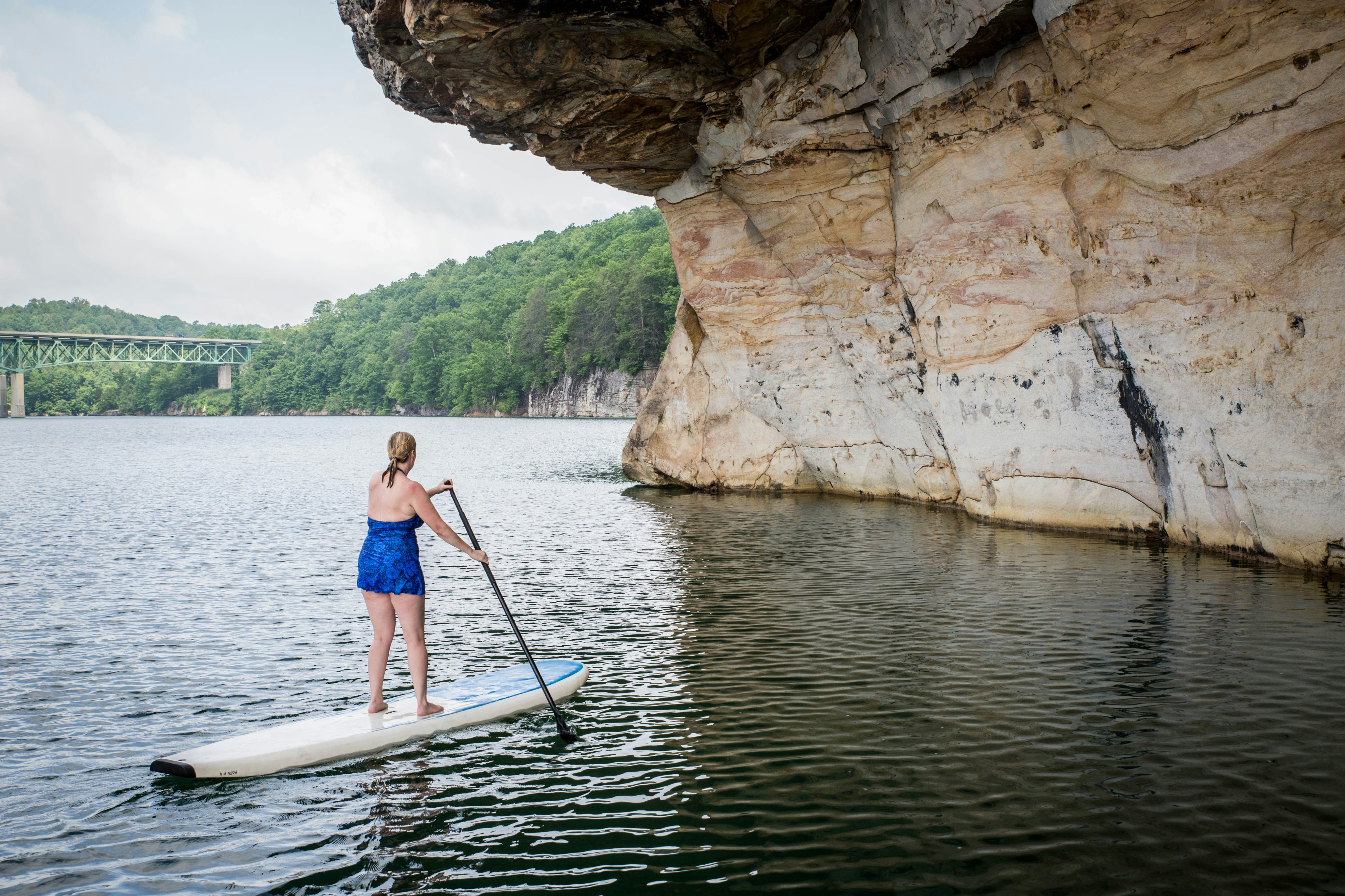 A woman paddleboards through calm waters at Summersville Lake, beneath a cliff face. To the left, the New River Gorge bridge is visible.