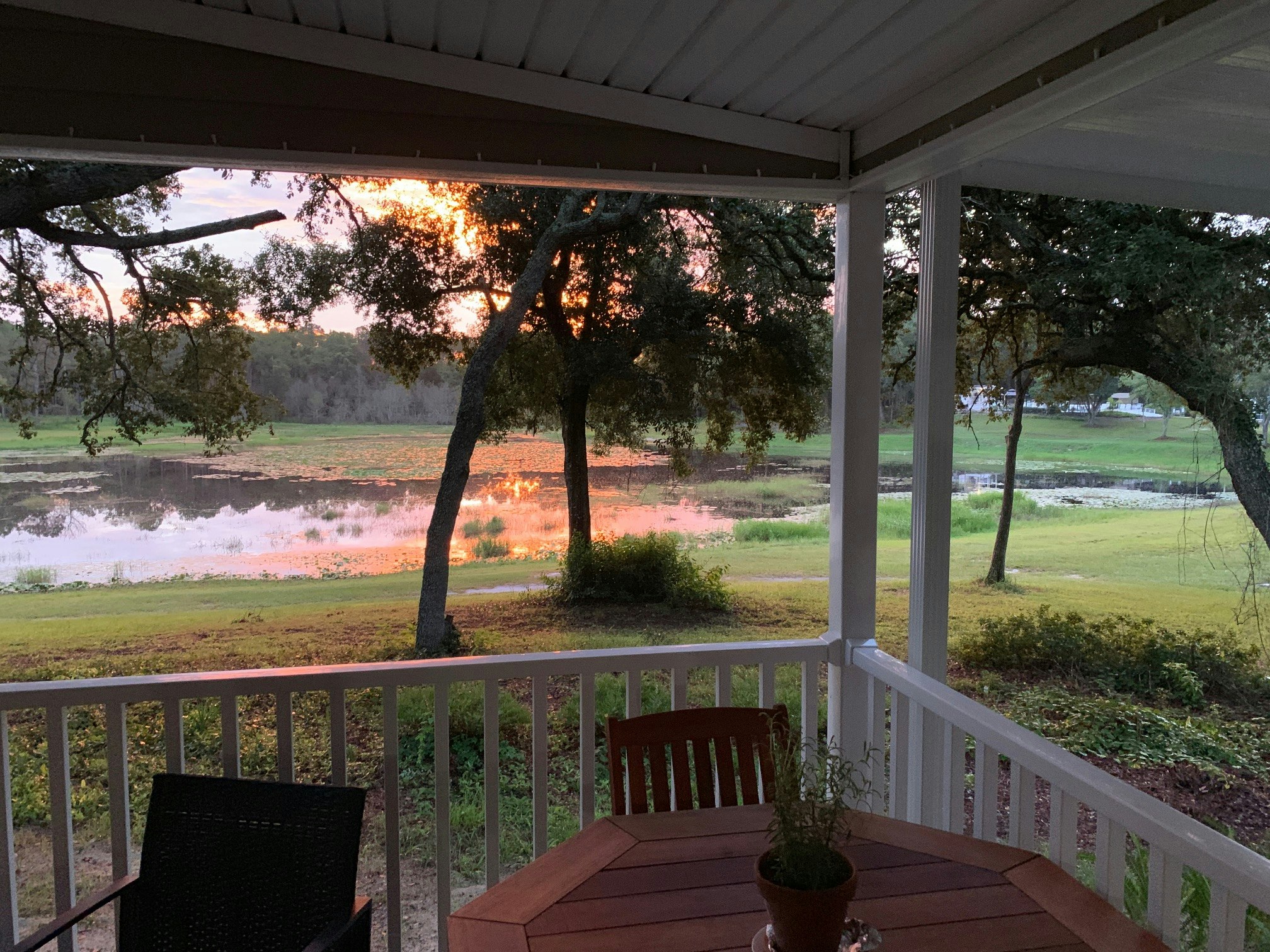 The corner of a whitewashed wooden porch furnished with a dark teak patio set frames a view of a small pond, which is reflecting a brilliant orange and pink Florida sunset at the Sunny Sands Naturalist Resort.