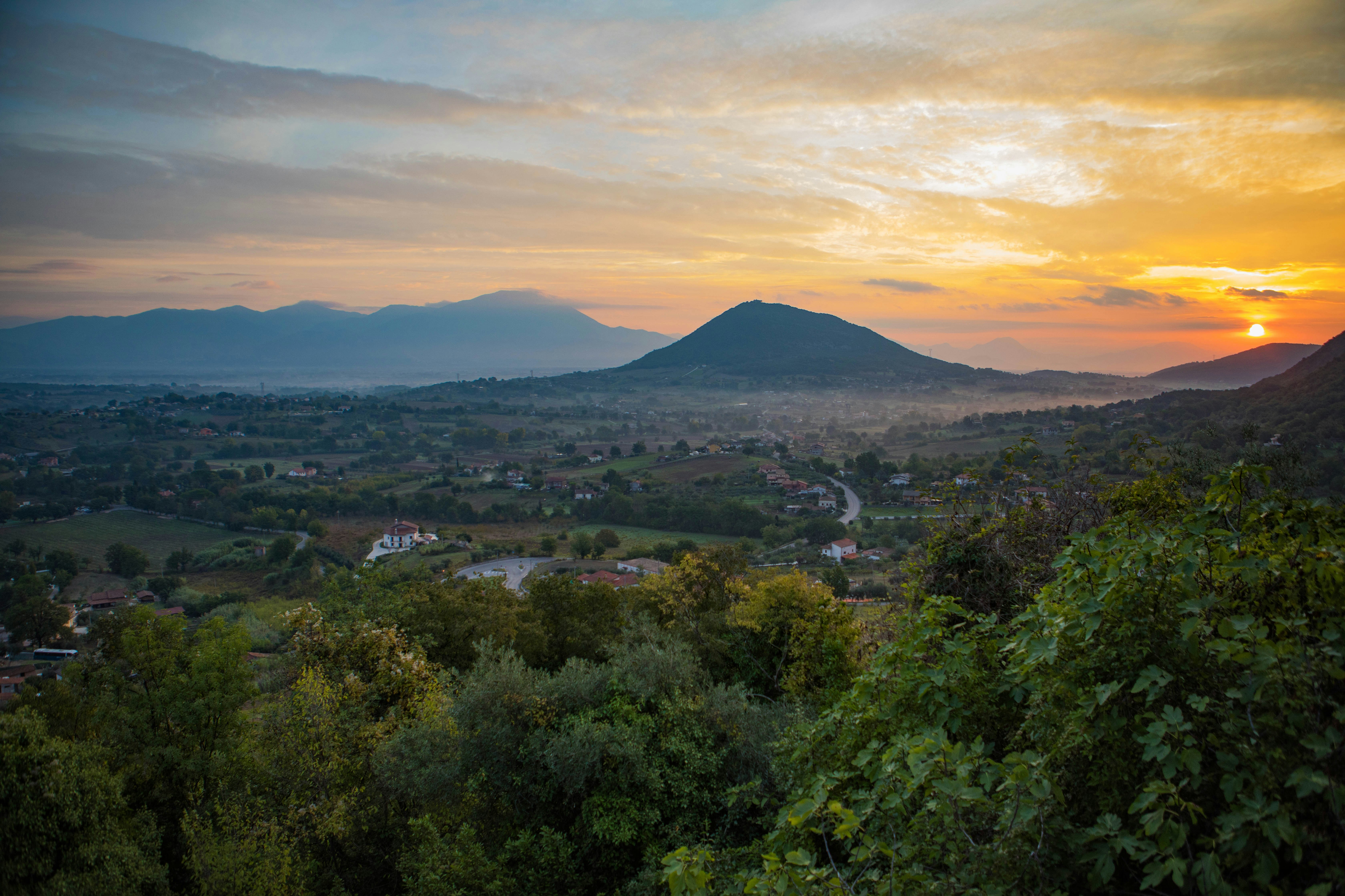 The sun rises over Pico, Italy with lush green foliage in the foreground, rolling hills dotted with Tuscan clay houses in the midground, and the striking blue mass of Pico mountain in the background surrounded by oranges and pinks in the sky. In the far distance is a blue, shadowed range of mountains separate from Pico