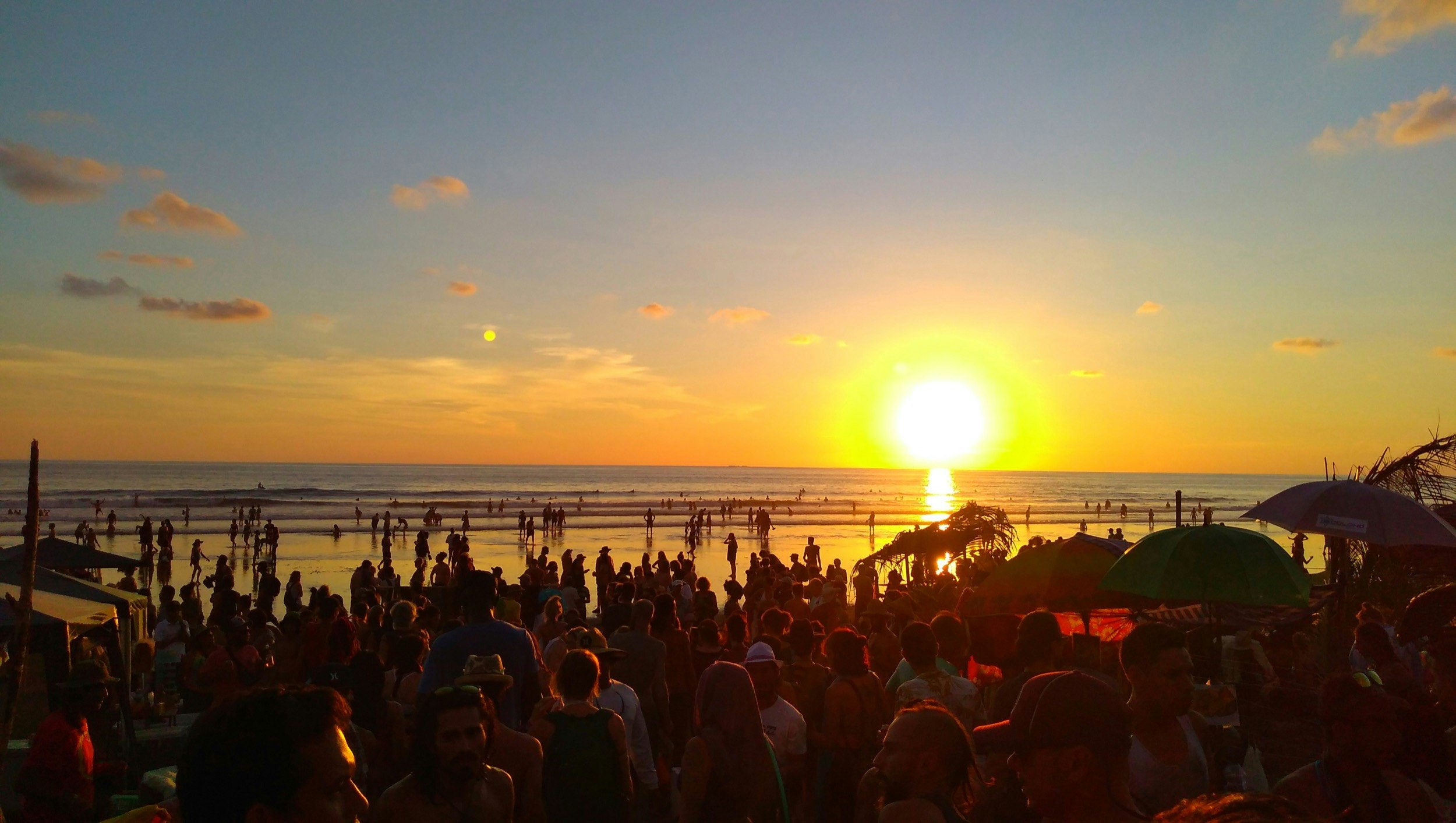 The sun sets over the pacific ocean as hundreds of people mill about on a Costa Rica beach and watch