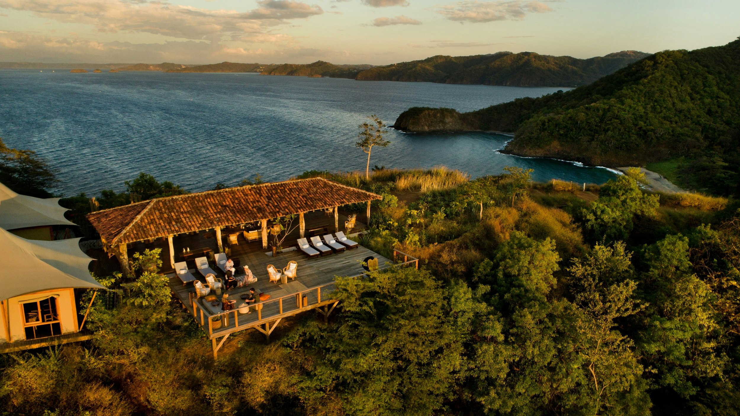 Aerial view of a wooden building near a beach on the edge of dense forest. The deck has several people on loungers or sat at the tables. Luxury tents are to the left