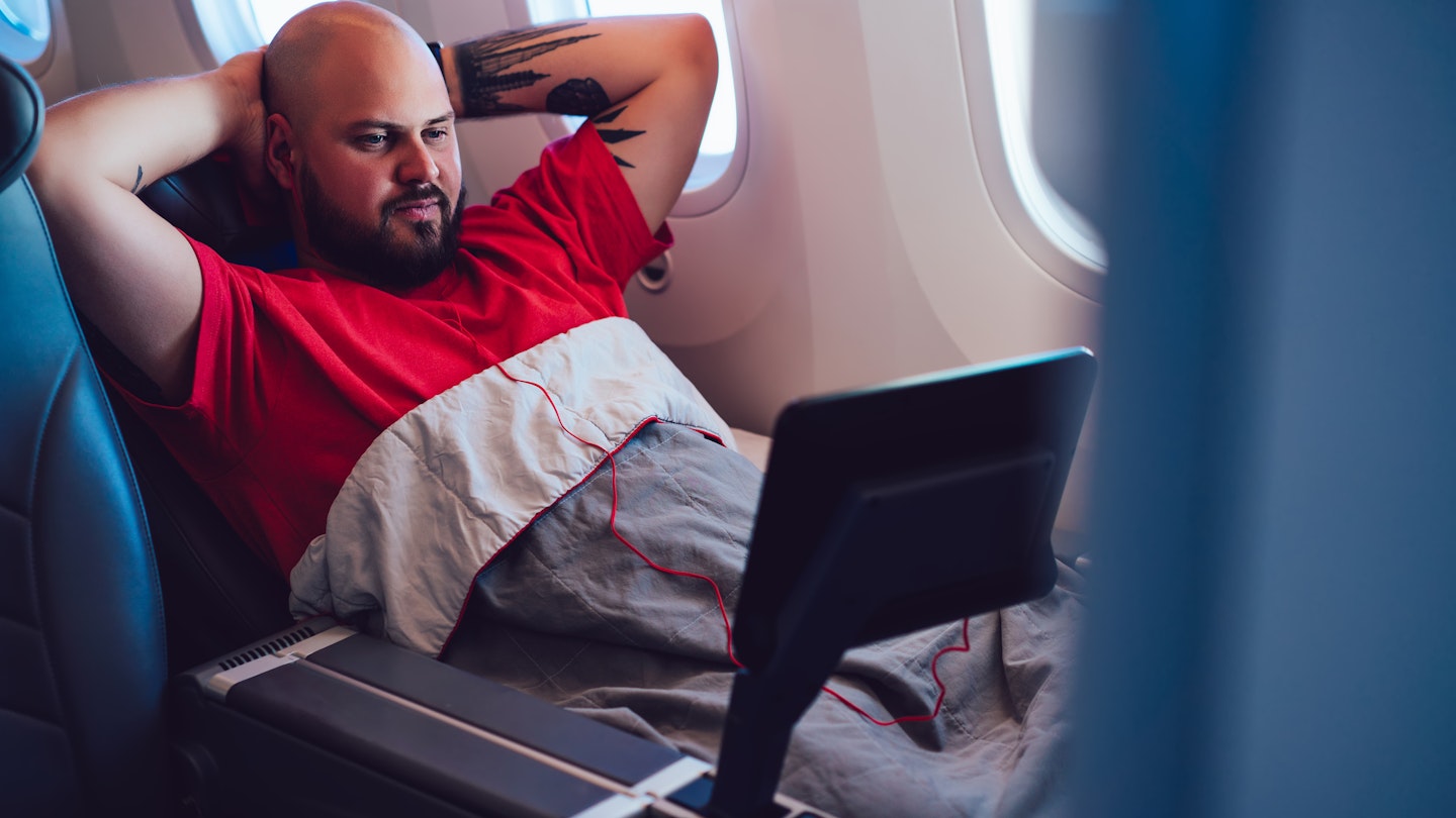A man reclines in an airplane seat with his hands folded behind his head as he watches a small screen. He's got a blanket pulled up to the middle of his chest and is next to the window. 