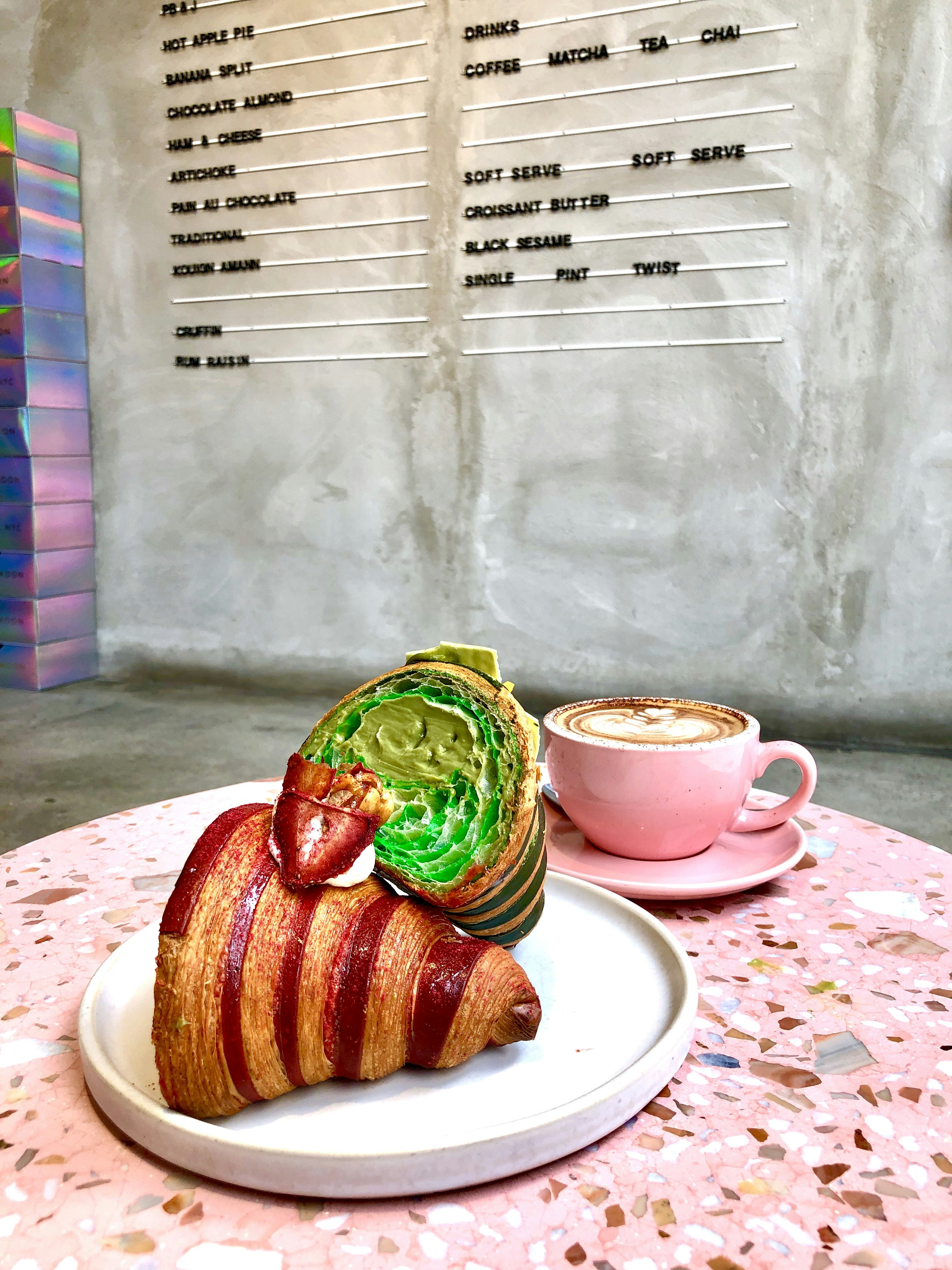 A croissant cut in half with green on the inside, garnished with a strawberry, beside a rose colored cup of coffee on a pink table and a menu on the wall behind it at Supermoon Bakehouse, New York City