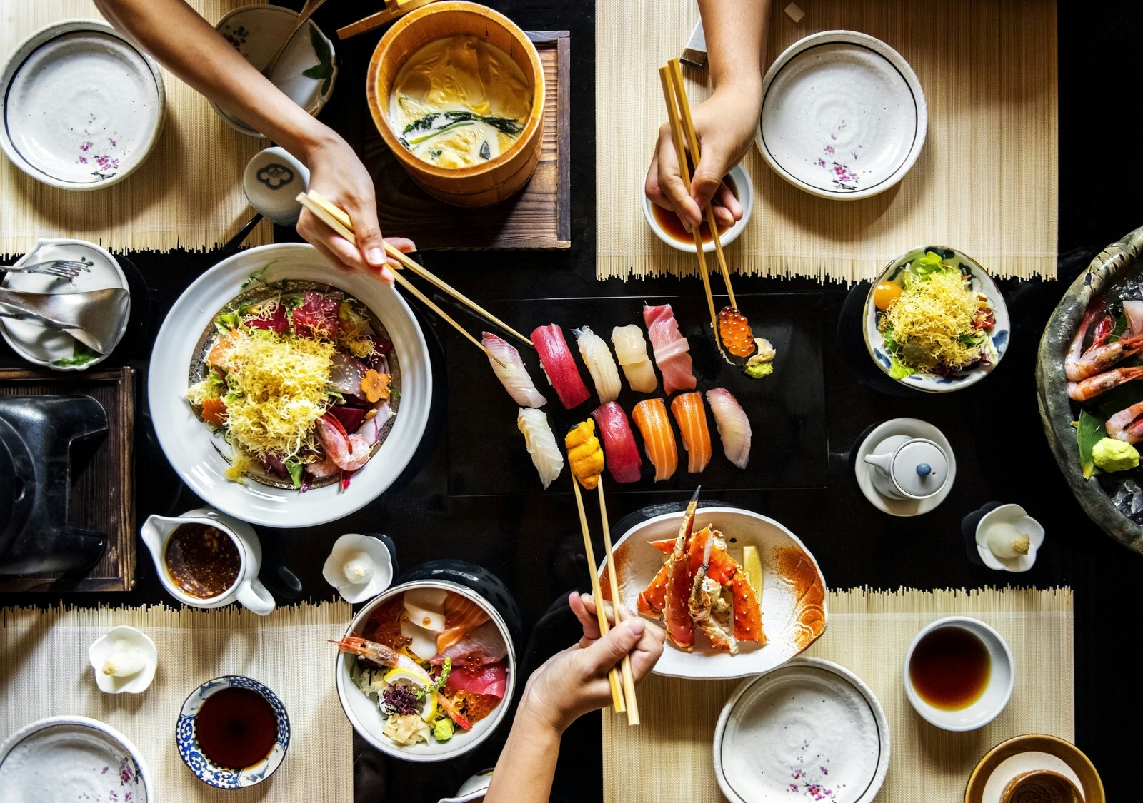 An aerial shot of a table with several sushi dishes laid out. There are three hands holding chopsticks reaching towards the dishes from around the table.