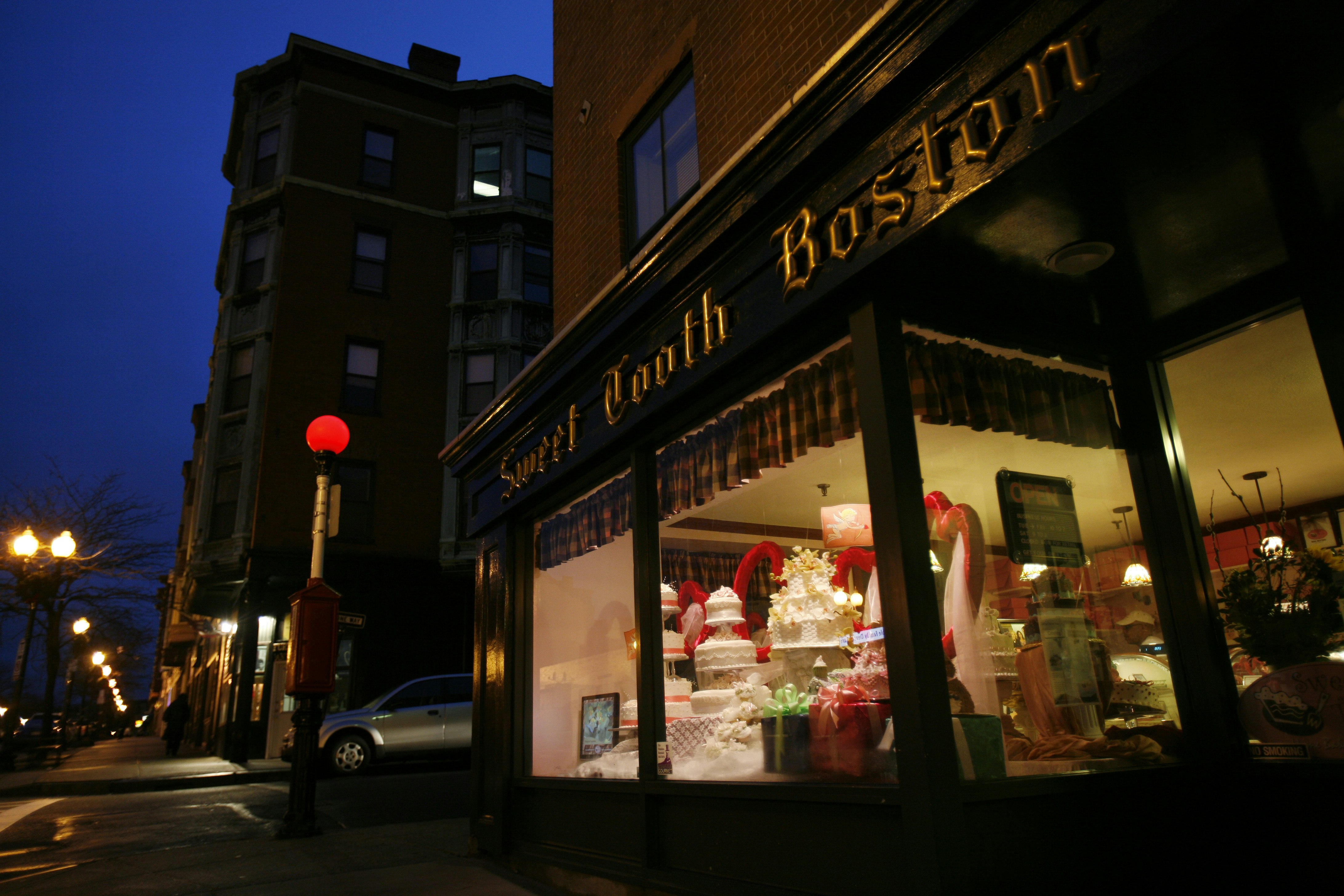 Exterior view of Sweet Tooth Boston at night. The golden letterings "Sweet Tooth Boston" catch the street lights. You can see large cakes and other decorations in the display window.