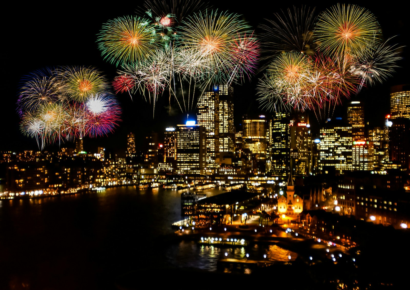 Sydney Harbour at nighttime with a colourful fireworks display happening over the water and skyscrapers