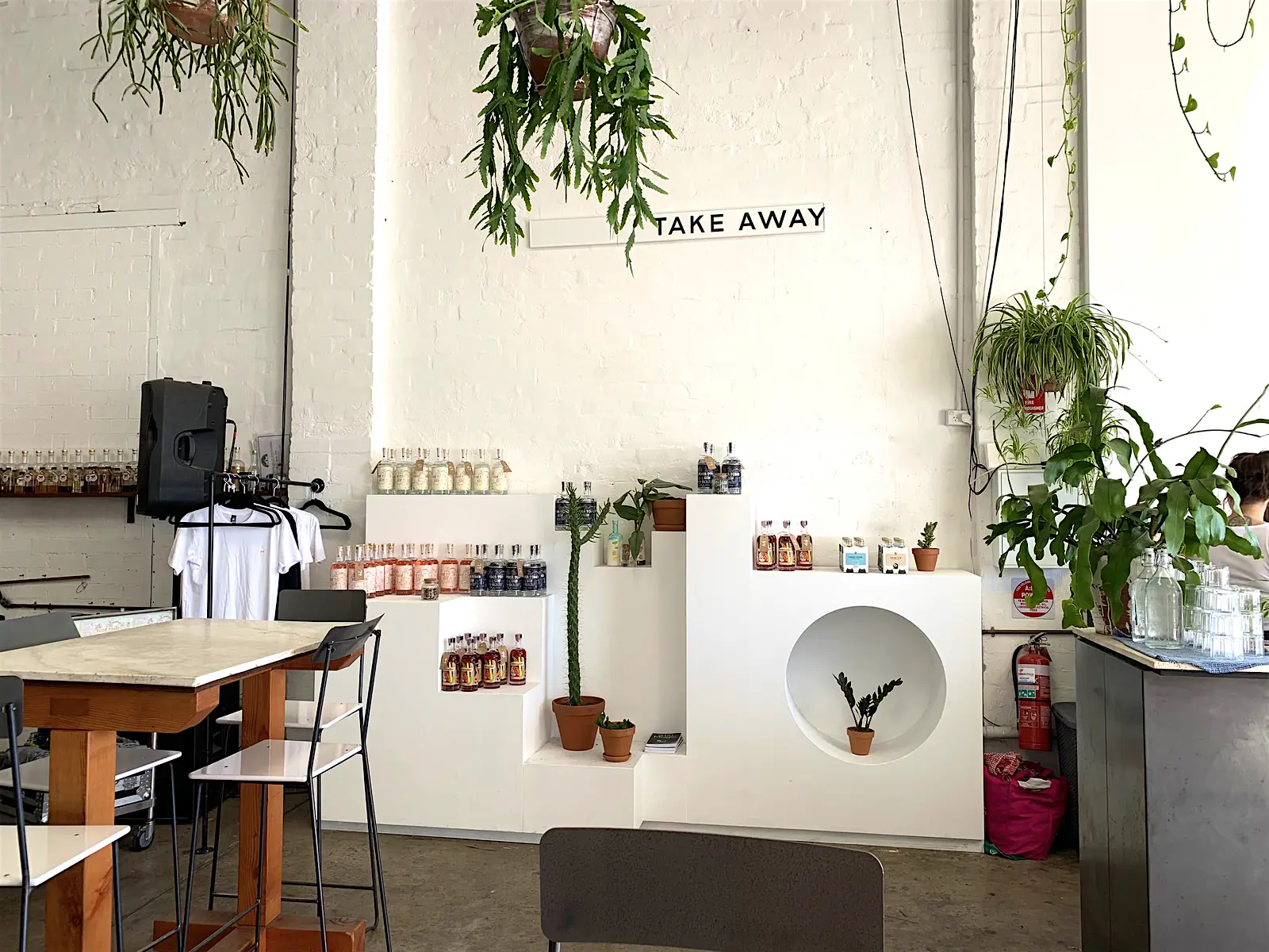 Sydney-gin-poor-toms-gin.jpg A brick wall painted white with a sign reading 'TAKE AWAY'. Underneath the sign different colour bottles of gin are arranged on an angular white built-in shelving unit along with potted plants.