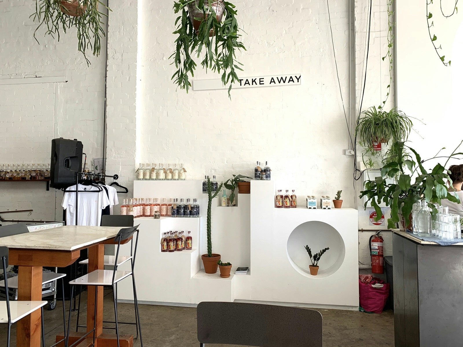A brick wall painted white with a sign reading 'TAKE AWAY'. Underneath the sign different colour bottles of gin are arranged on an angular white built-in shelving unit along with potted plants.