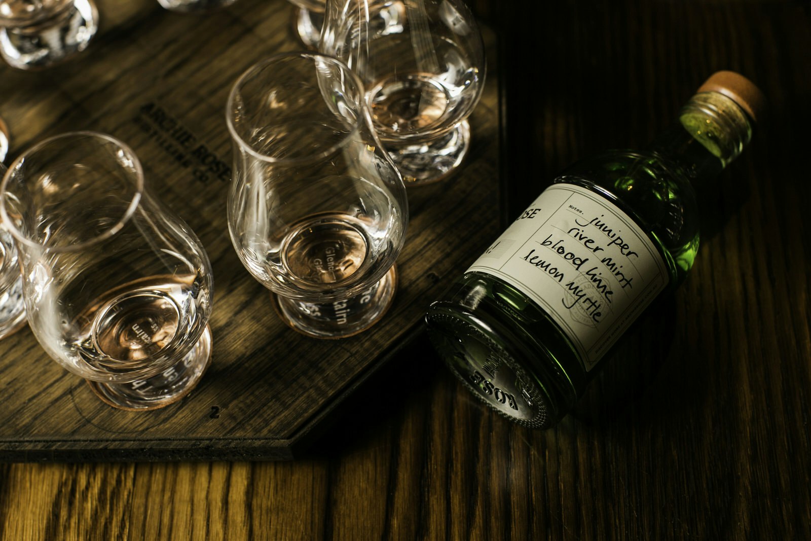 A small bottle of gin lies on its side on a wooden bar top next to several empty gin glasses. A hand-written label on the green bottle reads 'juniper river mint blood lime lemon myrtle'.