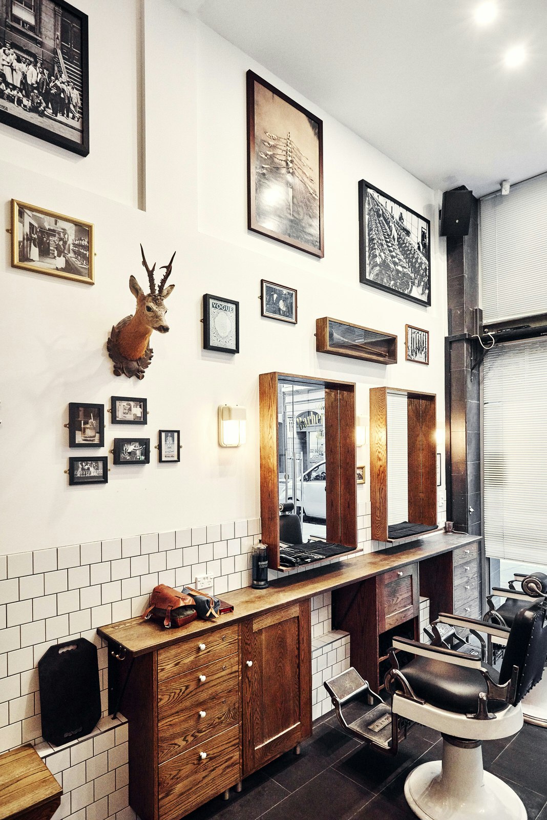 An old-fashioned barber shop interior with white tiled walls, framed photos, a small mounted deer's head and barbershop chairs