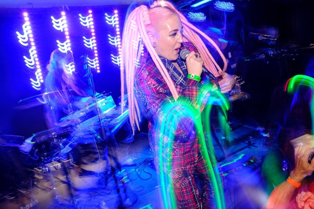 Performer Sykur hold a microphone to her mouth while dancing on stage. Her pink dreadlocked hair is bouncing around her face and she is on a multi-coloured stage at Airwaves Iceland music festival. 