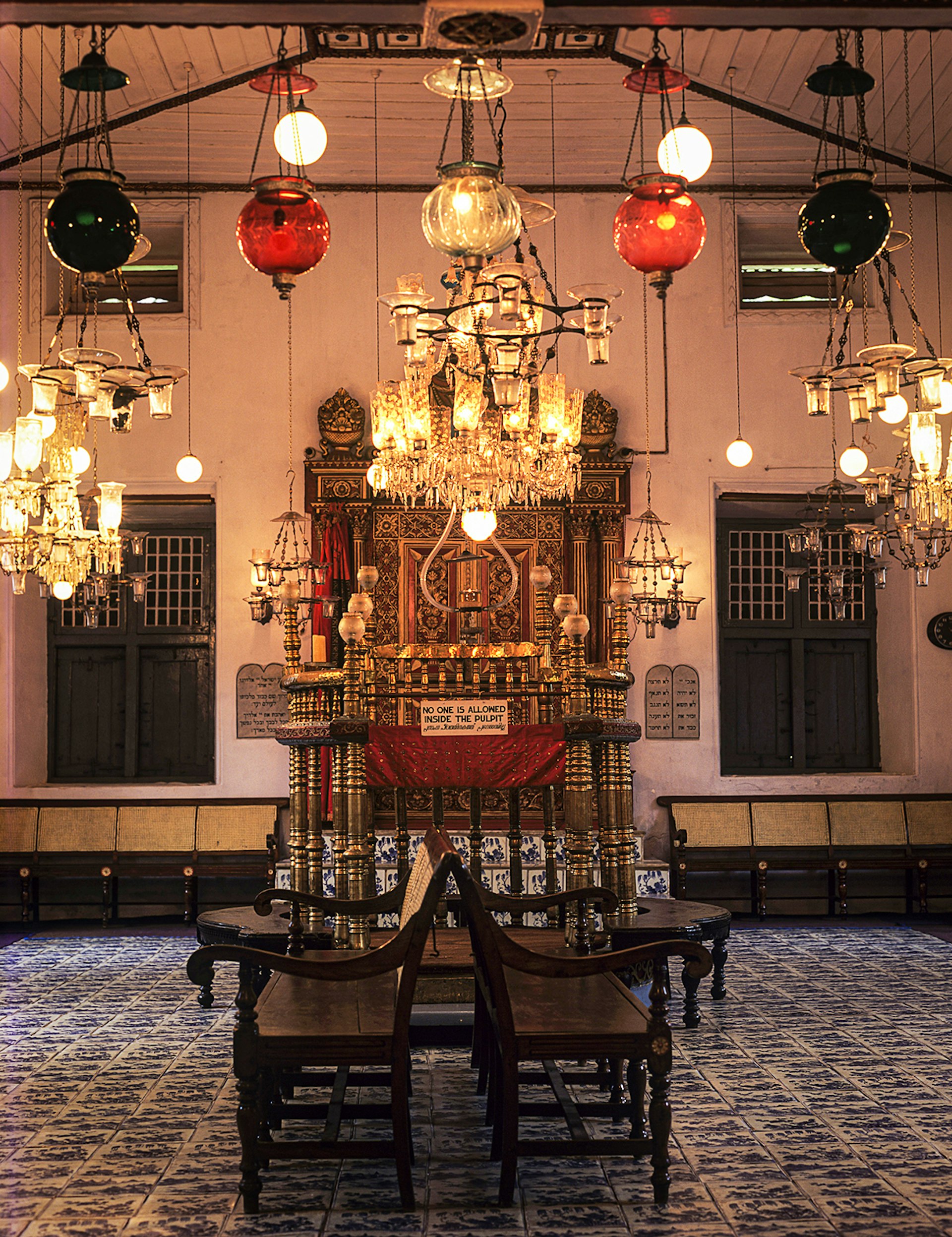 The interior of a synagogue featuring glass globe lanterns, chandeliers, and tiled floors. Kochi, India.