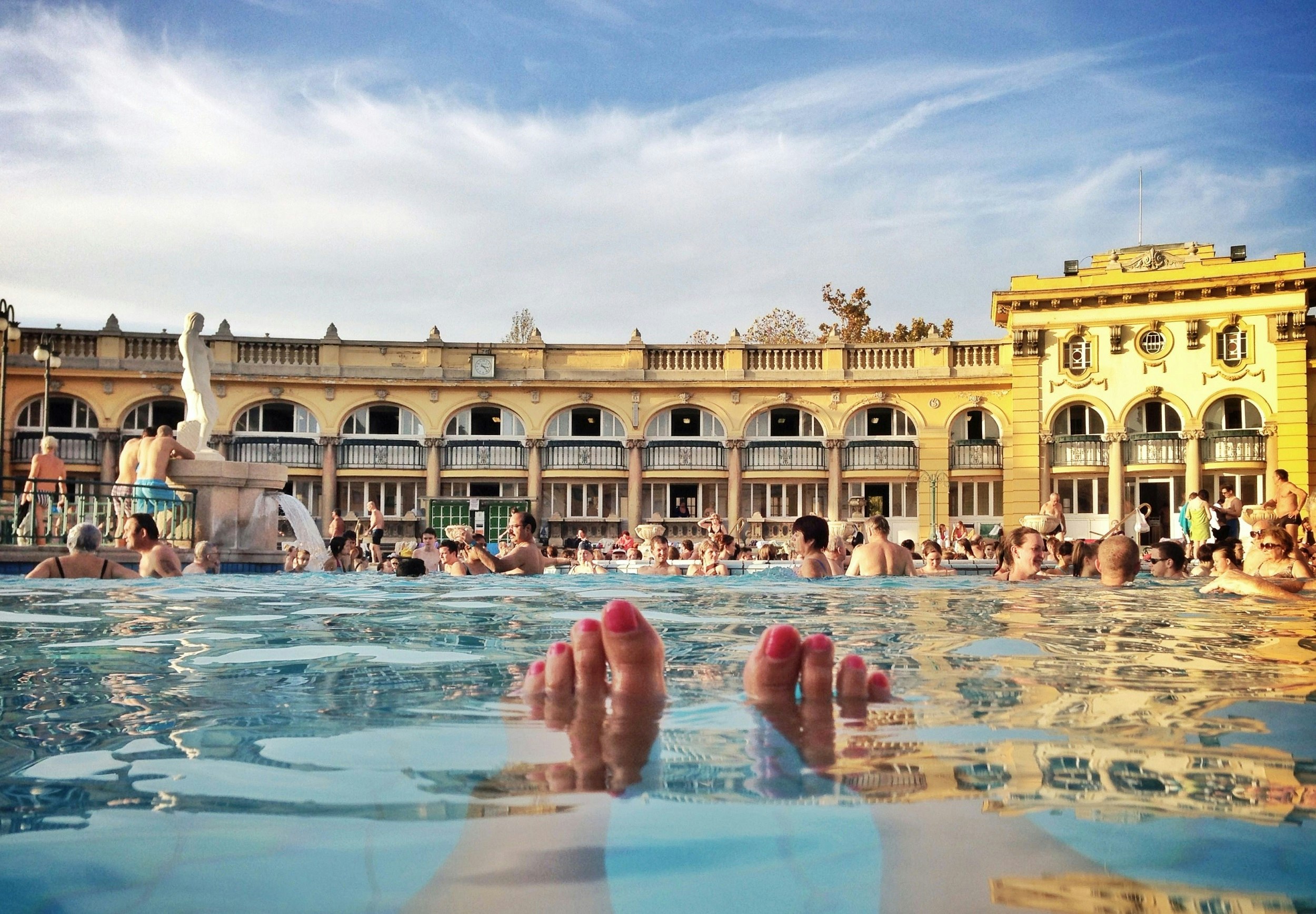 A point-of-view shot from someone in the outdoor pool at a thermal bath. Their feet are poking out of the water. Many other people relax in the pool. A curved yellow building with arched windows and balconies surrounds the thermal bath.