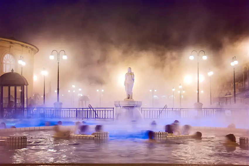 Szechenyi Baths at night in Budapest A nighttime shot of steam rising above a pool of water with several slightly blurred figures in it. There's a marble statue in the centre and several lit lampposts around the edge of the pool.