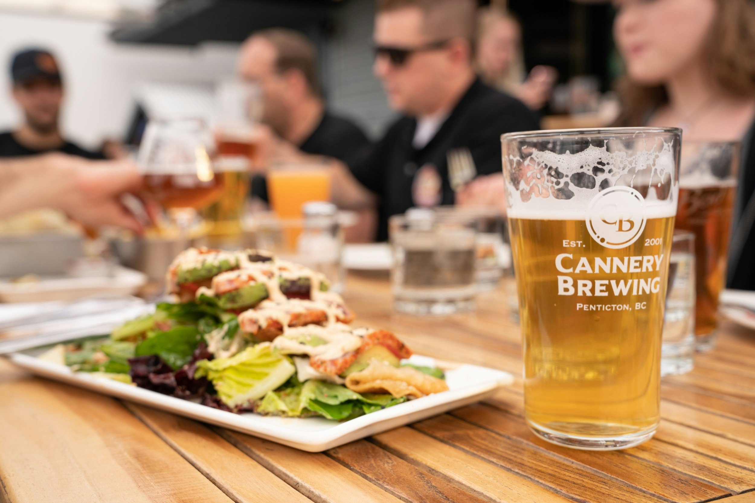 Ett glas ljust bärnstensfärgat öl ses i ett Cannery Brewing-glas, med en fräsch sallad och umgänge med människor i bakgrunden