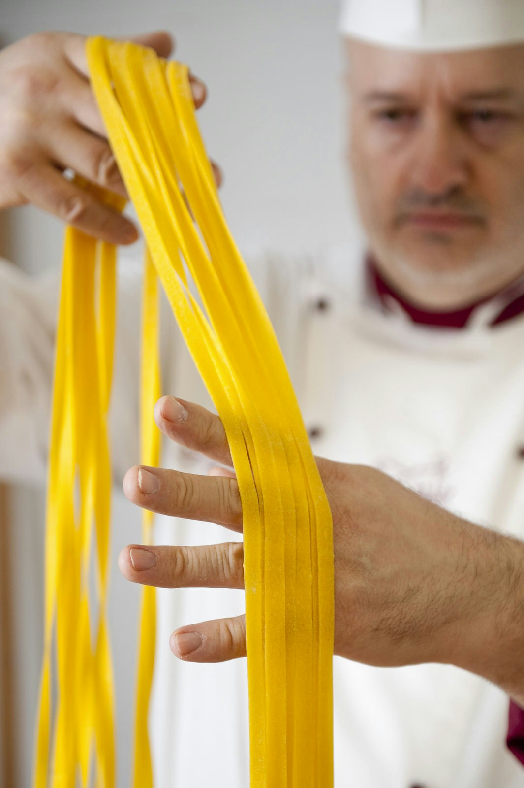 A close up of tagliatelle pasta being handled by chef Mauro Fabbri, who is slightly out of focus behind the tendrils of yellow pasta. 