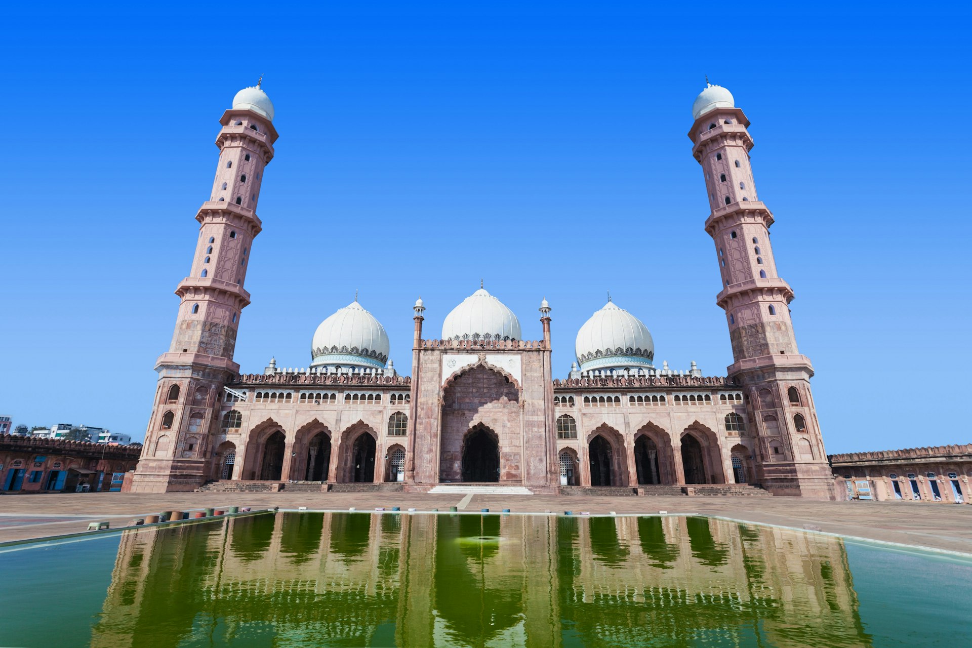 A view of Taj-ul-Masjid from the front, with the large mosque reflected in the pool used for washing in its central courtyard. The large mosque has three white domes and a towering minaret at both ends.