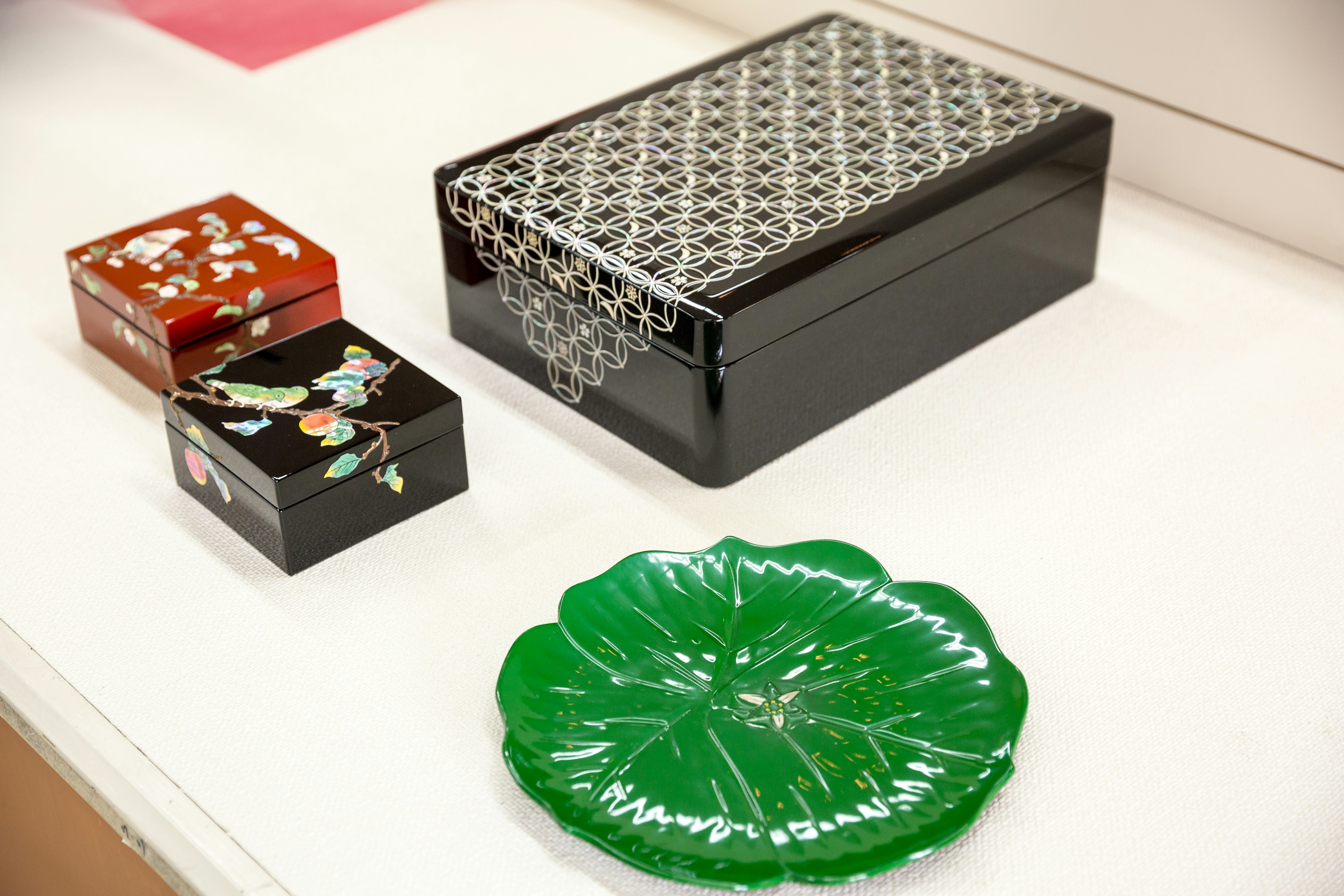 Two small jewellery boxes with intricate lacquerware birds and flowers on them, sat near a bento box with a geometric design, and a small green dish