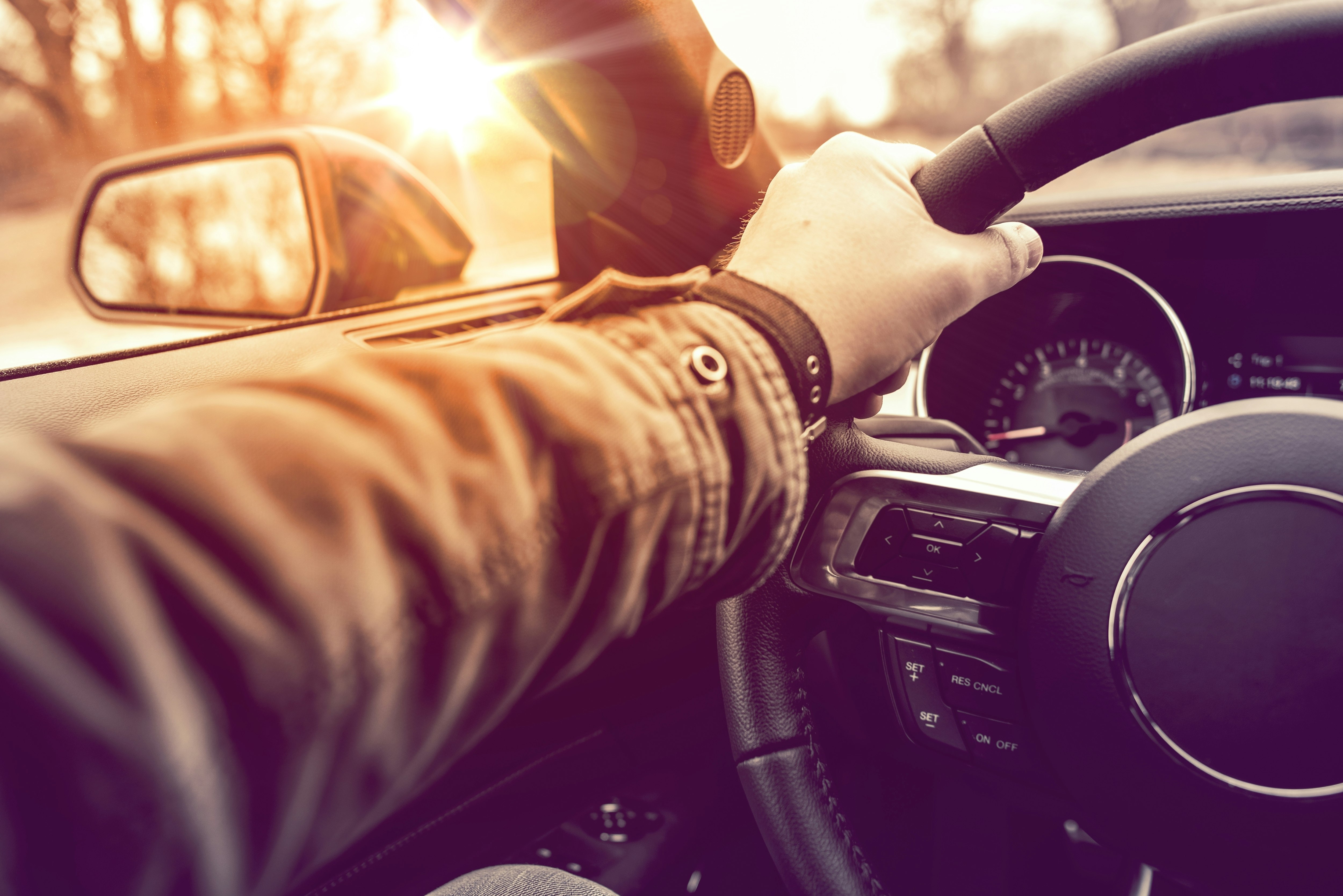 A man's hand is placed on a steering wheel in a car. The glare of the sun is visible through the drivers' seat window.