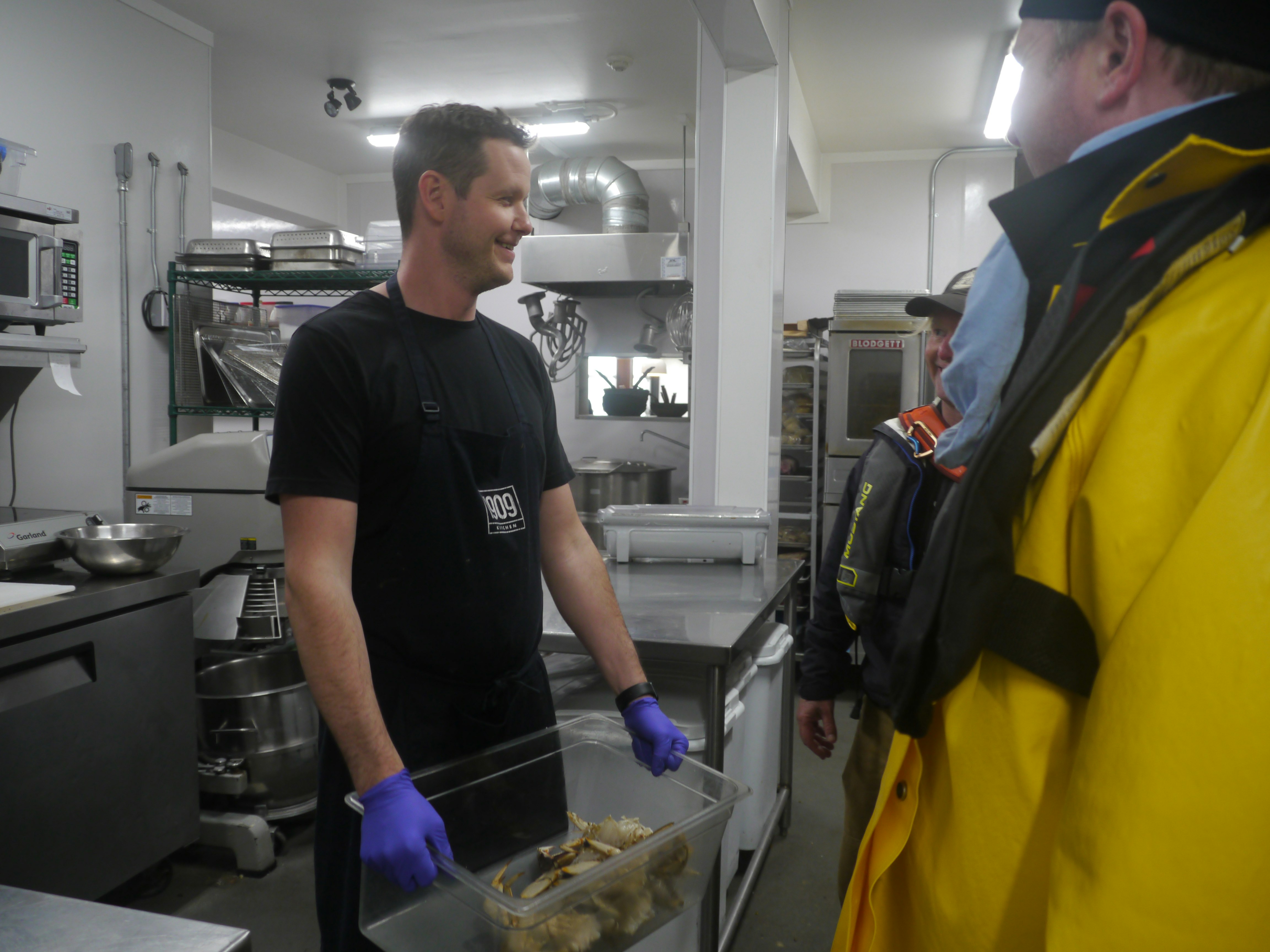 Chef Paul Moran stands back of house at 1909 Kitchen surrounded by stainless steel industrial cooking equipment and holding a plastic bin full of fresh crabs. He has blue plastic gloves on his hands and a big smile as he looks to the right side of the frame, where two fishermen are standing in yellow waterproof jackets.