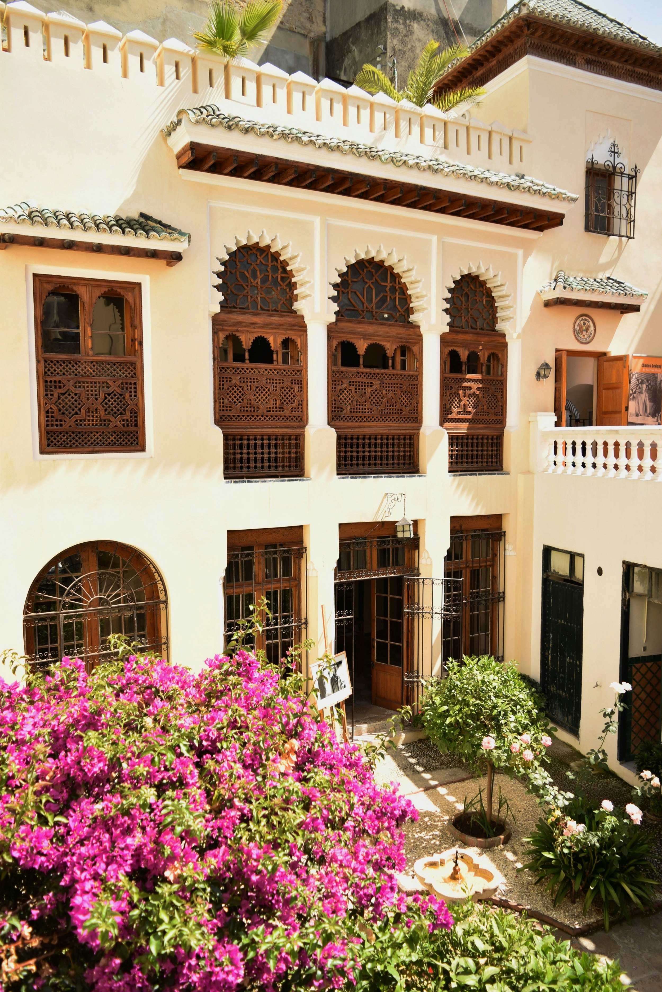 Tangier American Legation Museum; a courtyard view from the second level displaying a well manicured garden. The building features numerous ornate arches with delicately carved wooden lattices within the windows.