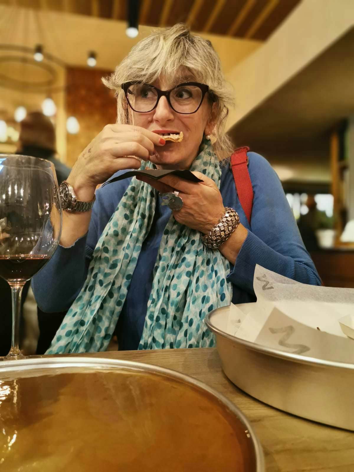 A woman taking a bite from a pastry held in her hand, using her other hand to hold a napkin close under her chin. She is looking away from the camera and there is a glass of red wine on the table in front of her
