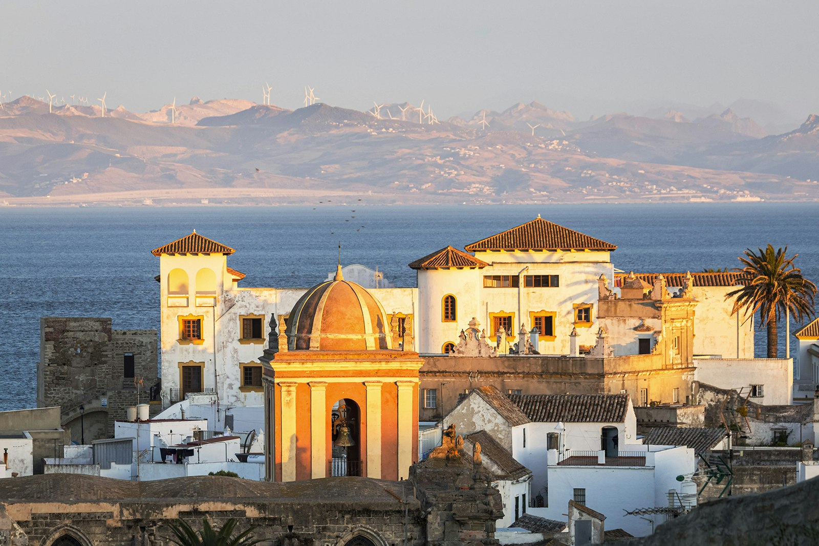 The historic center of Tarifa at sunset, with the ocean and a hill dotted with windfarms in the background. Cádiz, Spain.