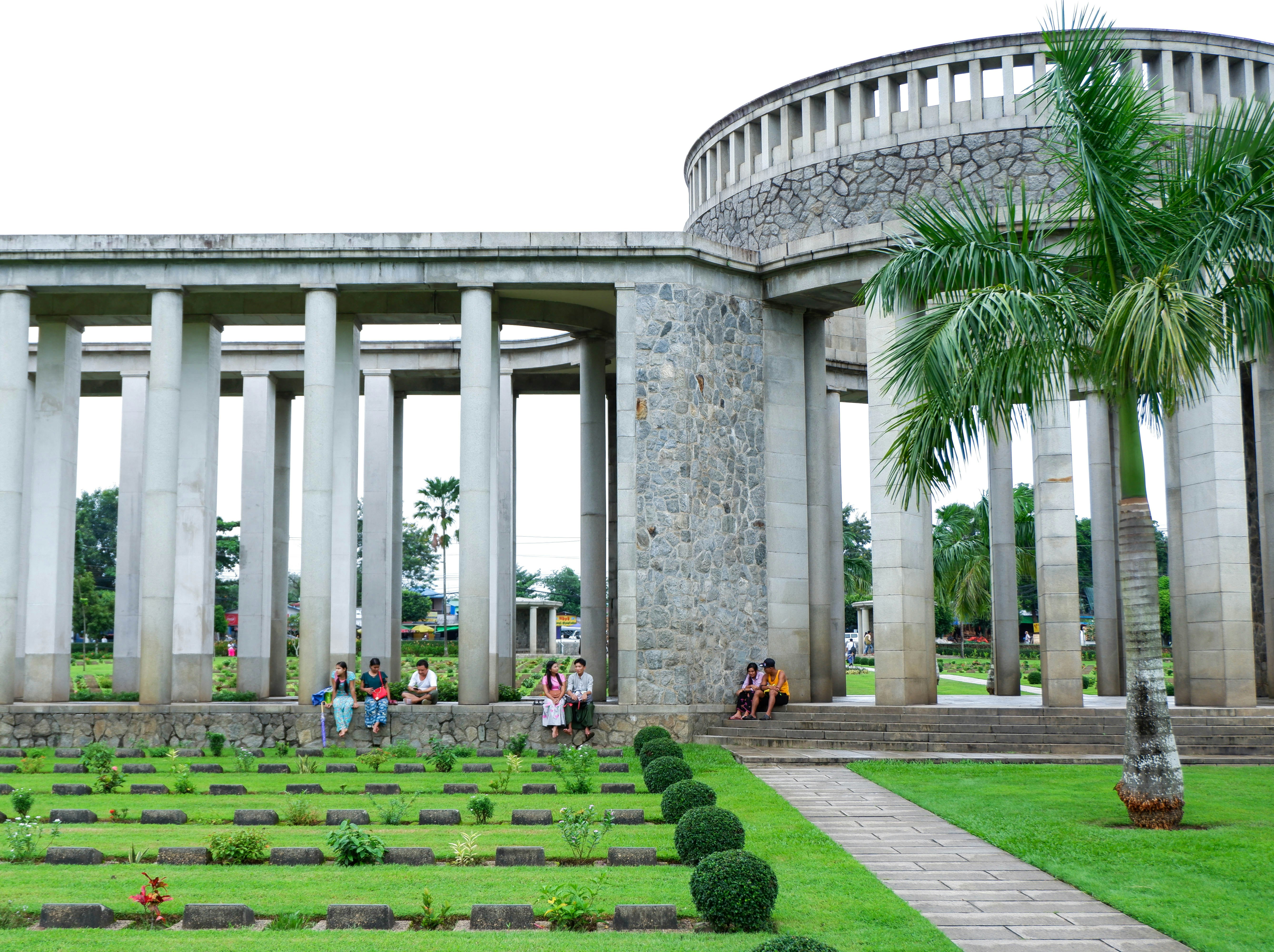 A few local people sit beneath the white stone pillars of Taukkyan War Cemetery's central monument. In the foreground numerous graves, with small headstones, are visible surrounded by manicured lawns.