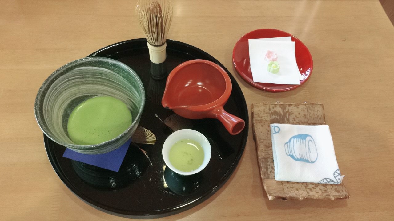 Matcha tea served at Takumi no Yakata on a black tray. The tea is in a small green bowl, alongside a red pourer, wooden whisk and a small cup