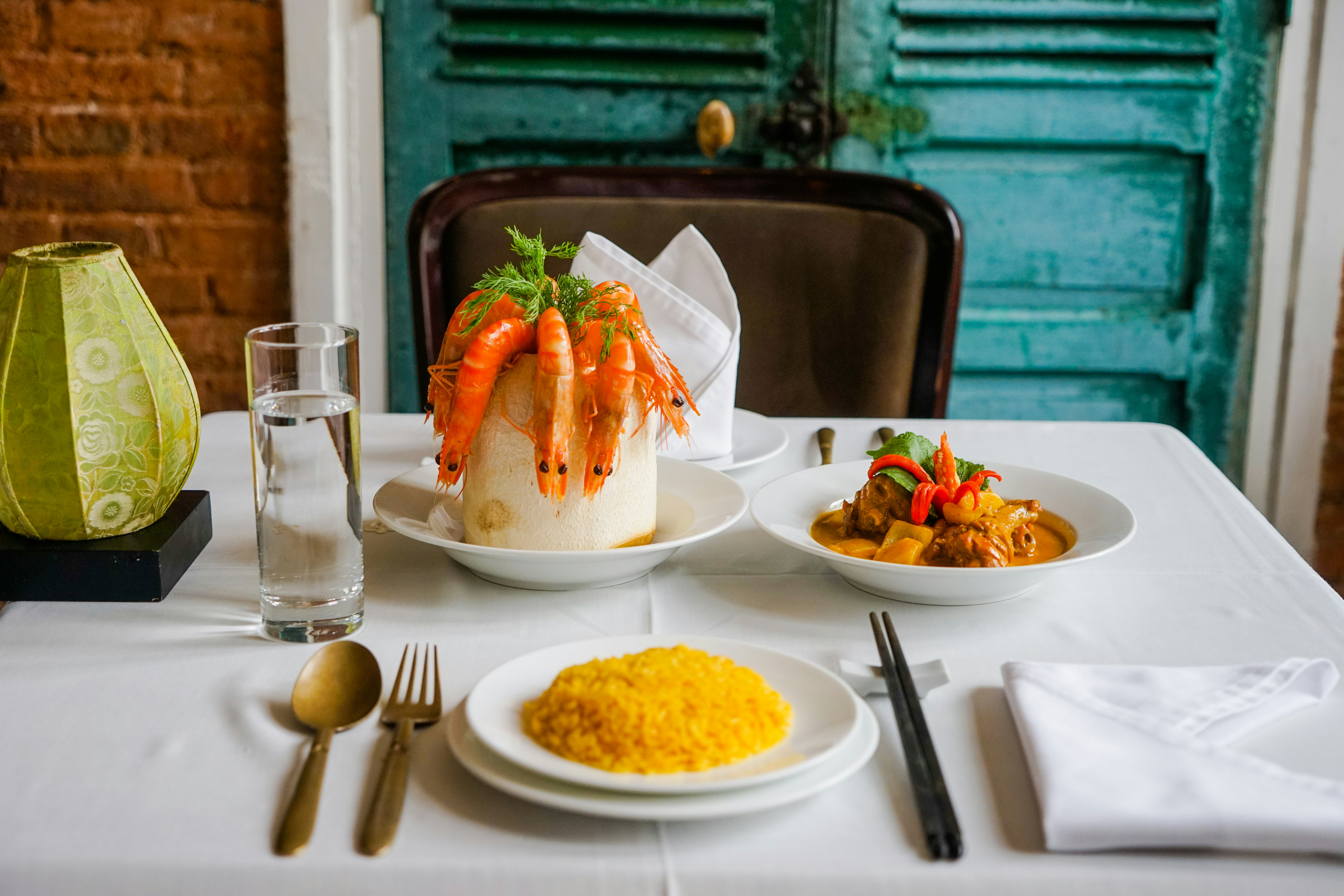 A table is set with dishes, cutlery and a glass of water. One dish is filled with yellow rice, another filled with vegetables and meat in an orange sauce and third has cooked shrimp dangling from a circular dish. 