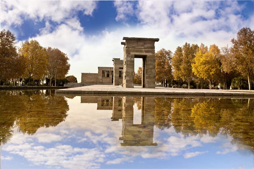 Three stone arches of Templo de Debod on a stone platform over a reflecting pool surrounded by trees with yellowing leaves in Madrid