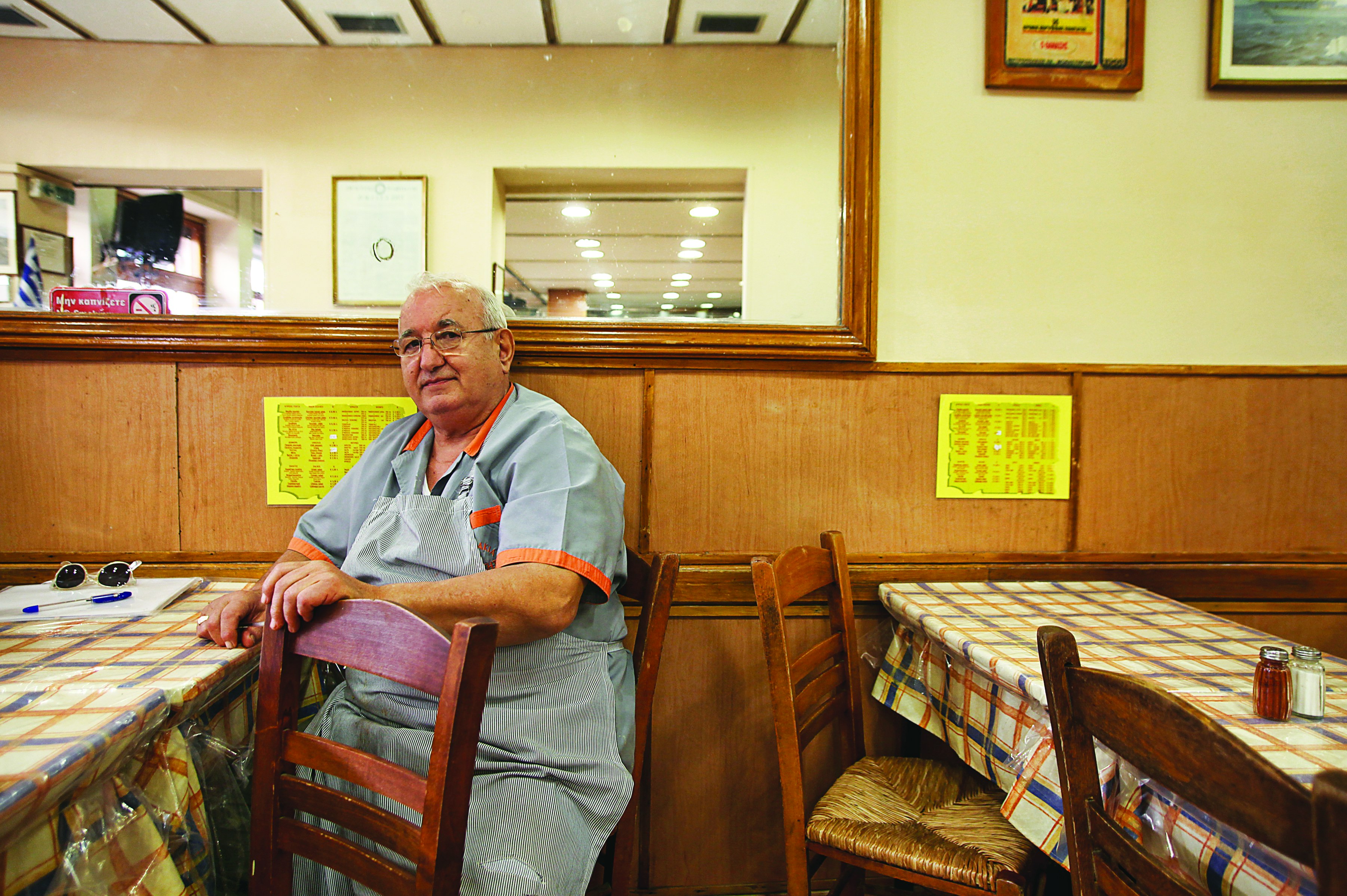 The owner of Thanasis restaurant sits at one of the tables, whcih are covered in blue and orange plaid clothes covered in clear plastic. He is wearing a light blue chefs shirt with orange trim on the sleeves and collar. The walls are paneled in wood and veneer, belaying the age of the restaurant