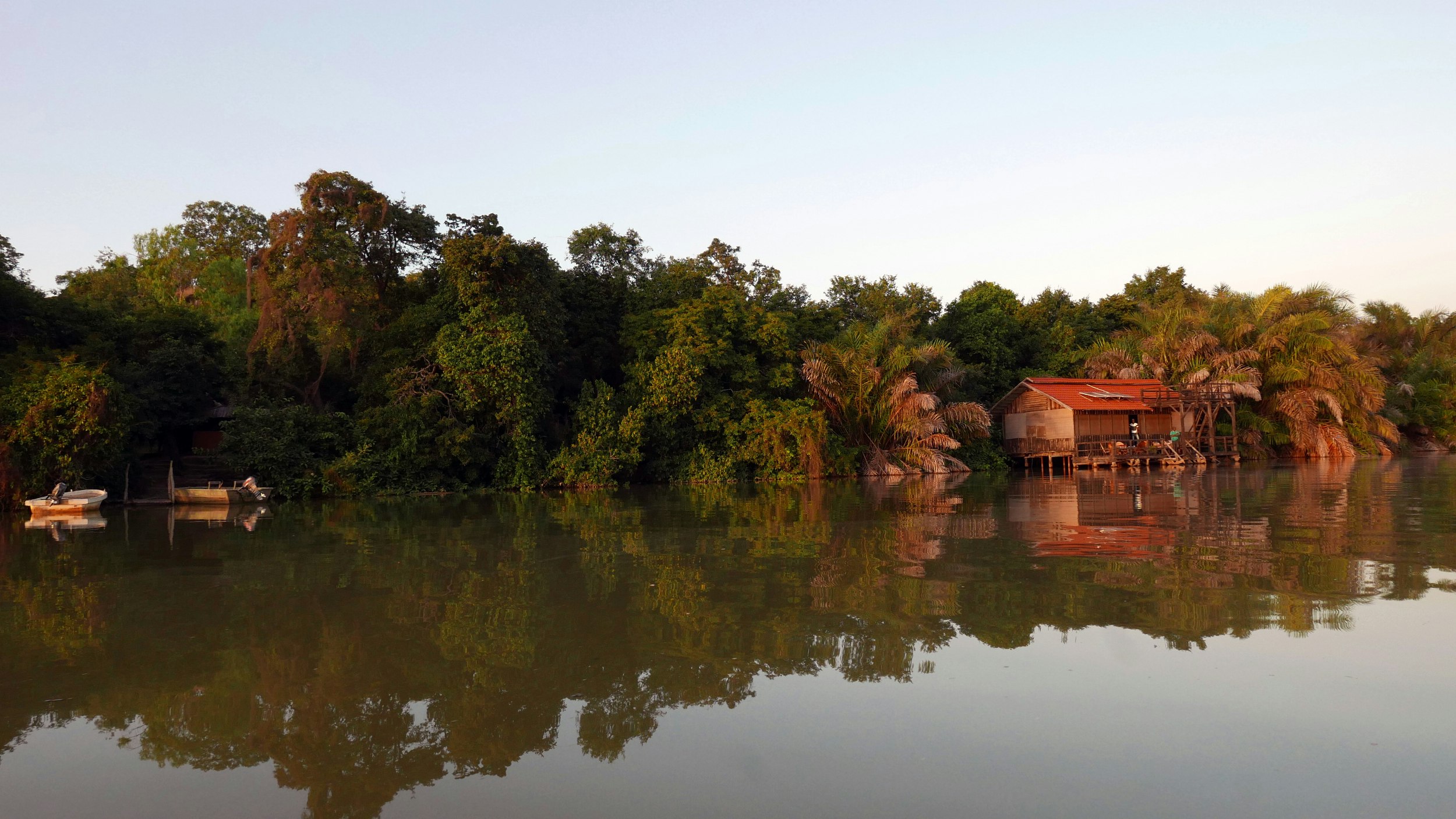 A forested section of riverbank reflects in the mirror-like surface of the River Gambia; nestled into the trees is a red-tinned roof wooden building - nearby are a couple of small boats moored on shore.