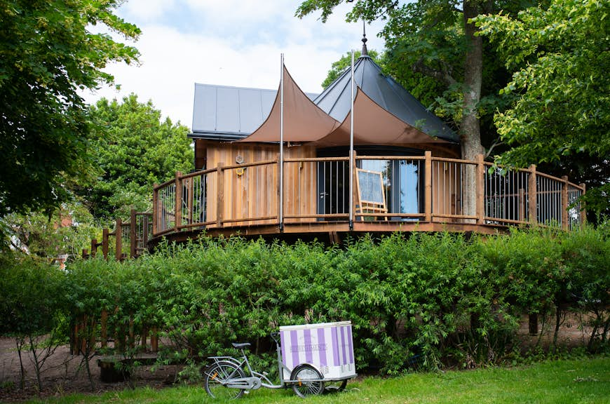 A large treehouse overlooks a garden in the English countryside. Green trees are visible in the background.