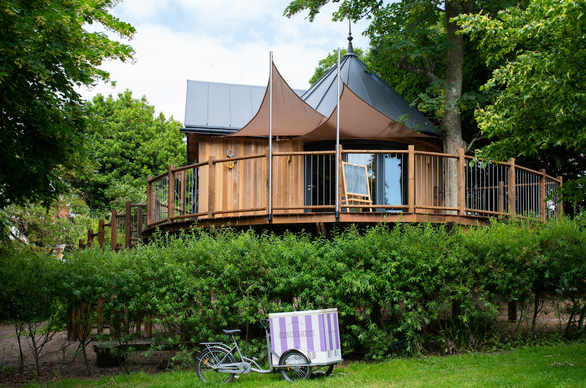 A large treehouse overlooks a garden in the English countryside. Green trees are visible in the background.