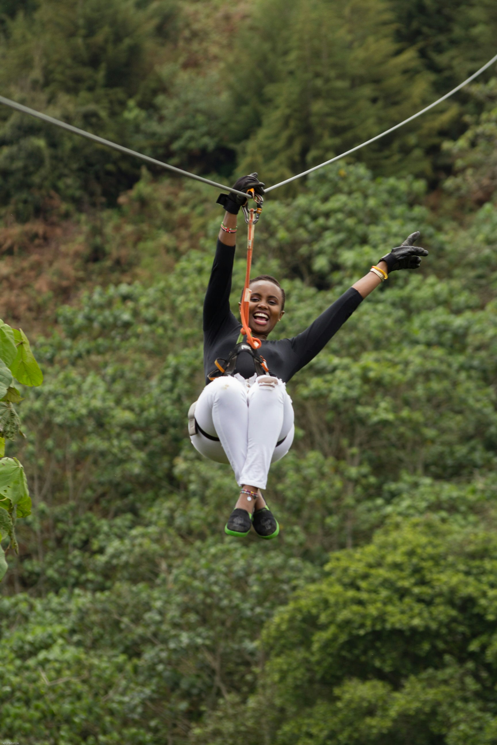 A woman smiles widely as she careers down a zip wire through the forest with one arm in the air, joyfully.