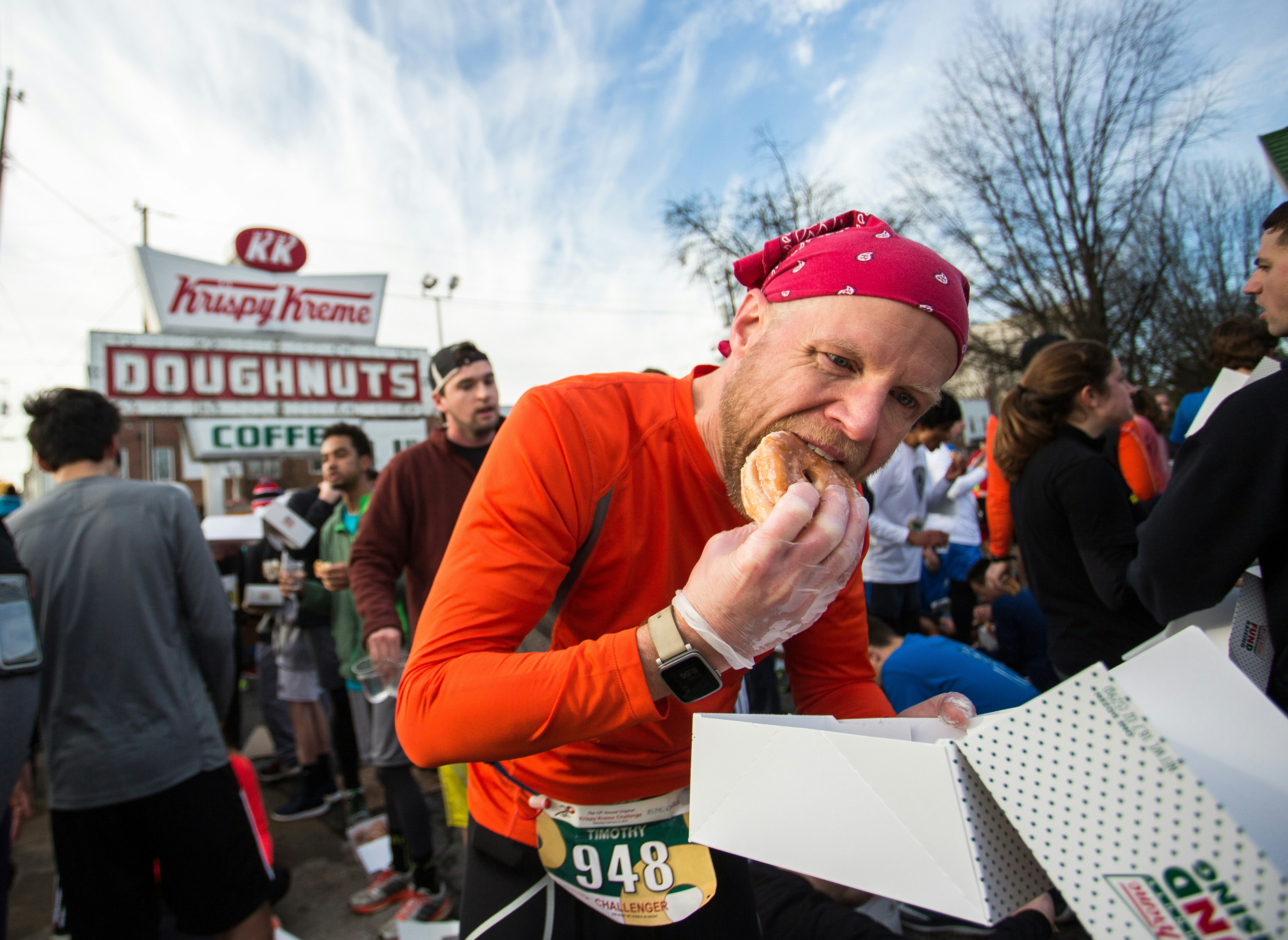 A man shoves a doughnut into his mouth during a run. In the background is a Krispy Kreme stand  