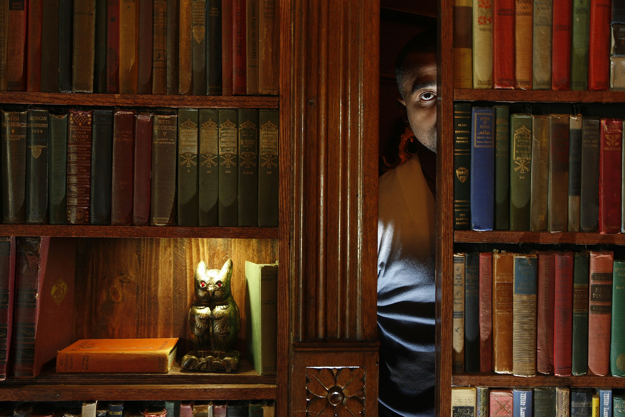 A man peeks from behind a secret door behind a bookcase. On a shelf is a golden owl statue below a bright light.