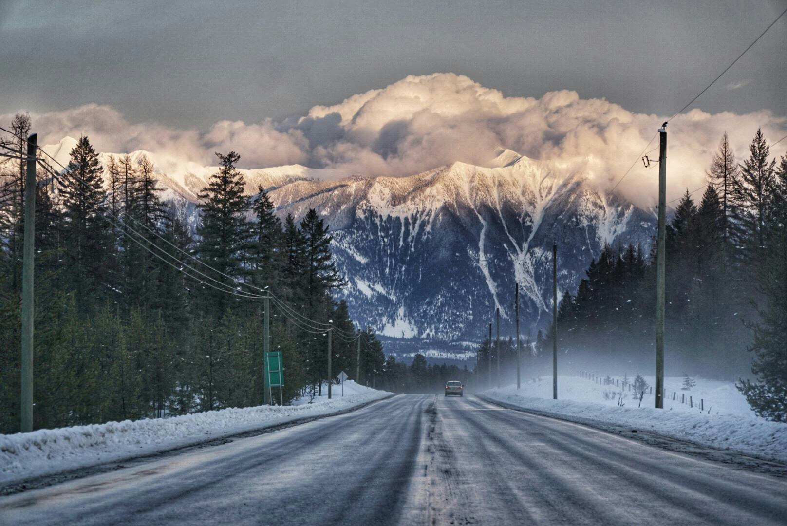 A vehicle is in the middle distance, driving along a snowy road lined with tall pine trees. Snow-laden, forested mountains are straight ahead, and huge clouds linger on top of them, under a grey sky.